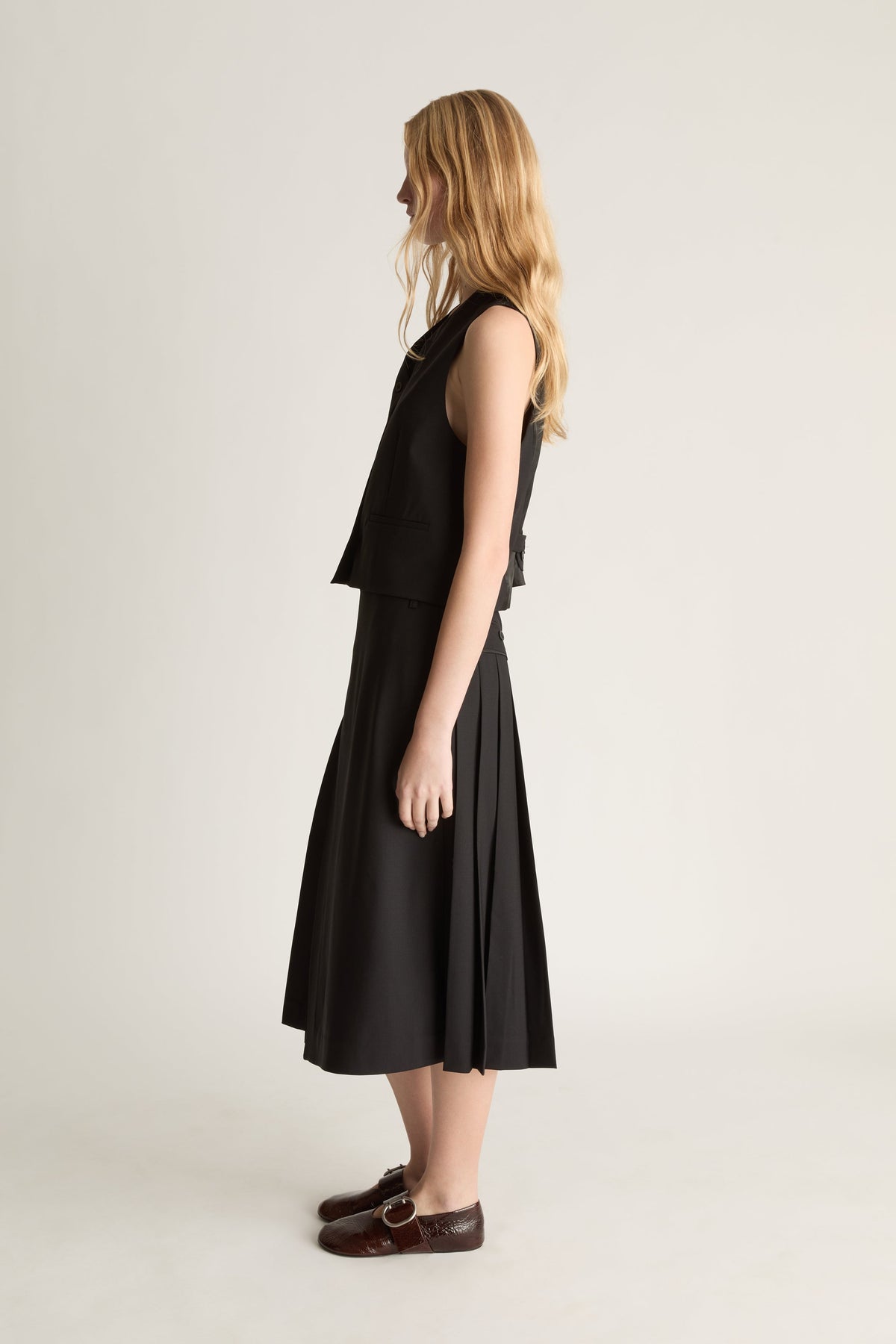 Side view of a woman wearing a black vest and a black kilt style wool skirt against a white studio background. 