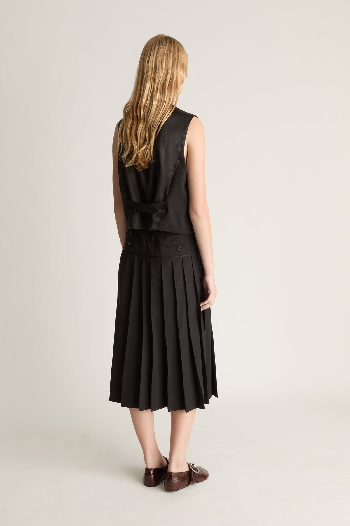 Back view of a woman wearing a black sleeveless top and pleated skirt on a plain background