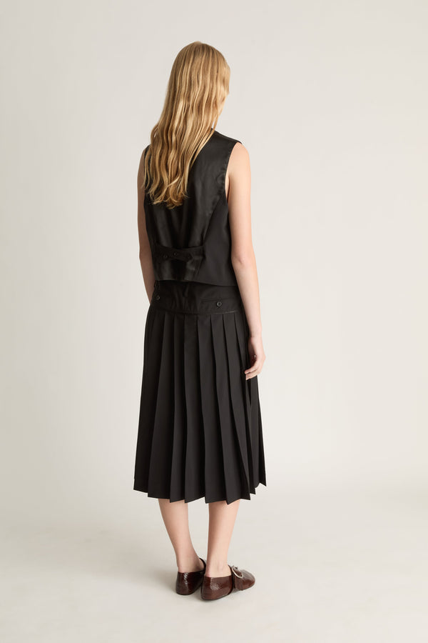 Back view of a woman wearing a black sleeveless top and pleated skirt on a plain background