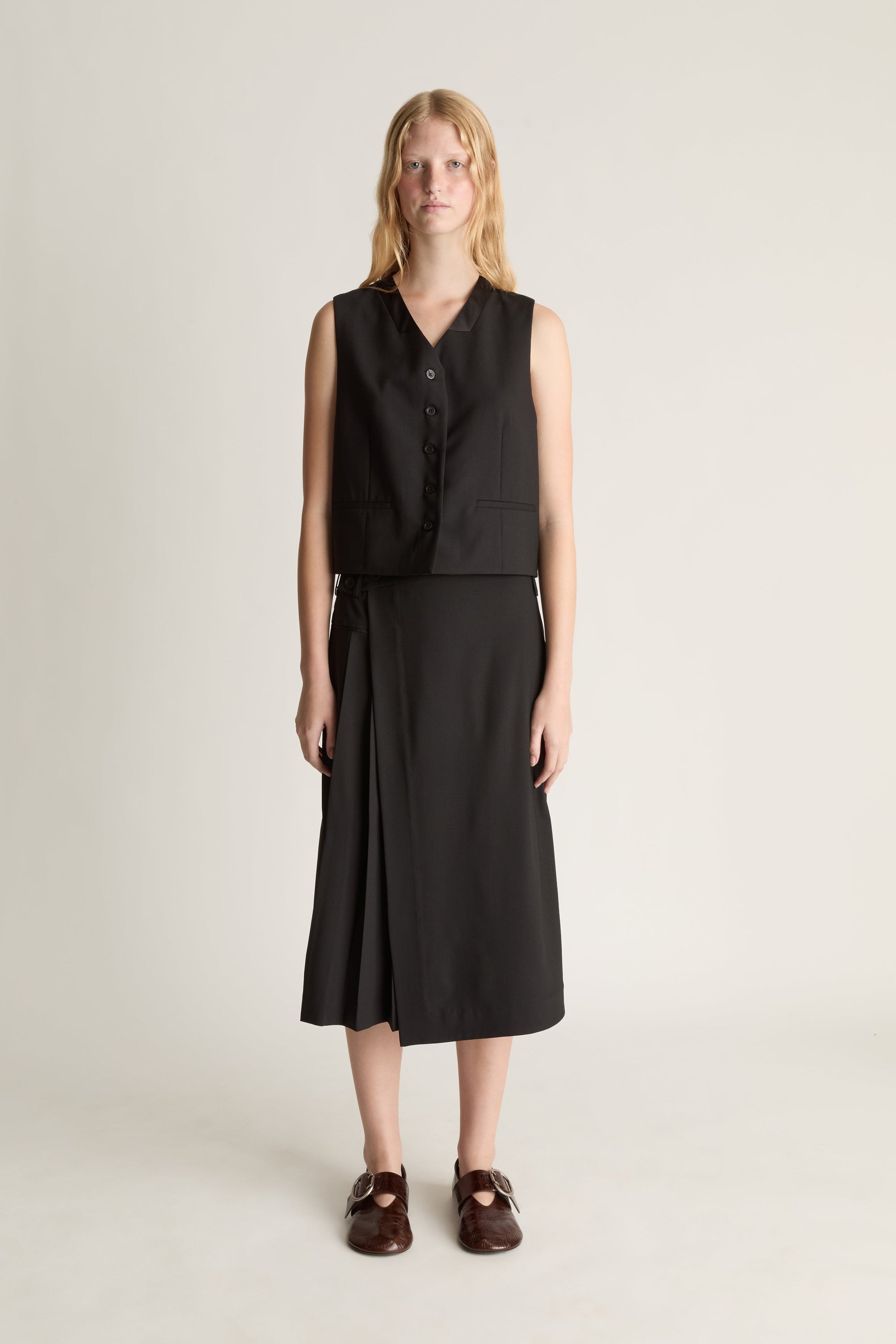 A woman stands against a plain light beige studio background wearing a black sleeveless button-front vest over a matching black midi skirt, with dark brown loafers.
