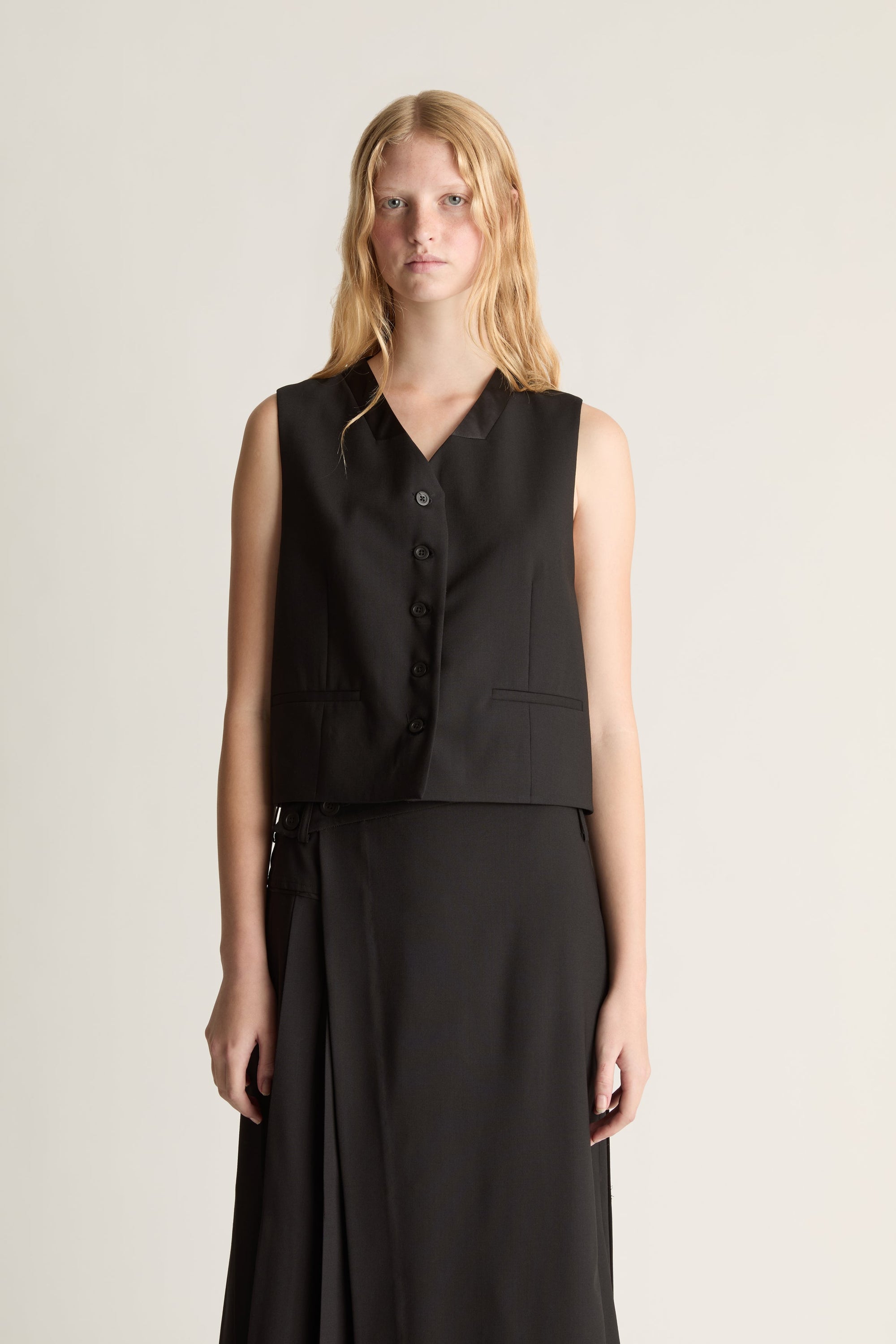 A close-up of a model wearing a sleeveless black vest with a button front and a matching black skirt, standing against a plain light beige studio background.