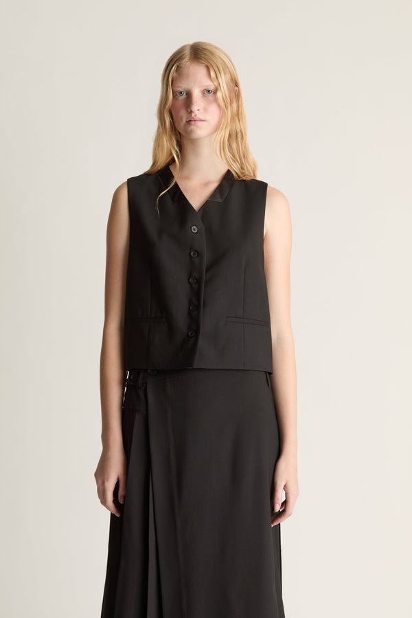 A close-up of a model wearing a sleeveless black vest with a button front and a matching black skirt, standing against a plain light beige studio background.