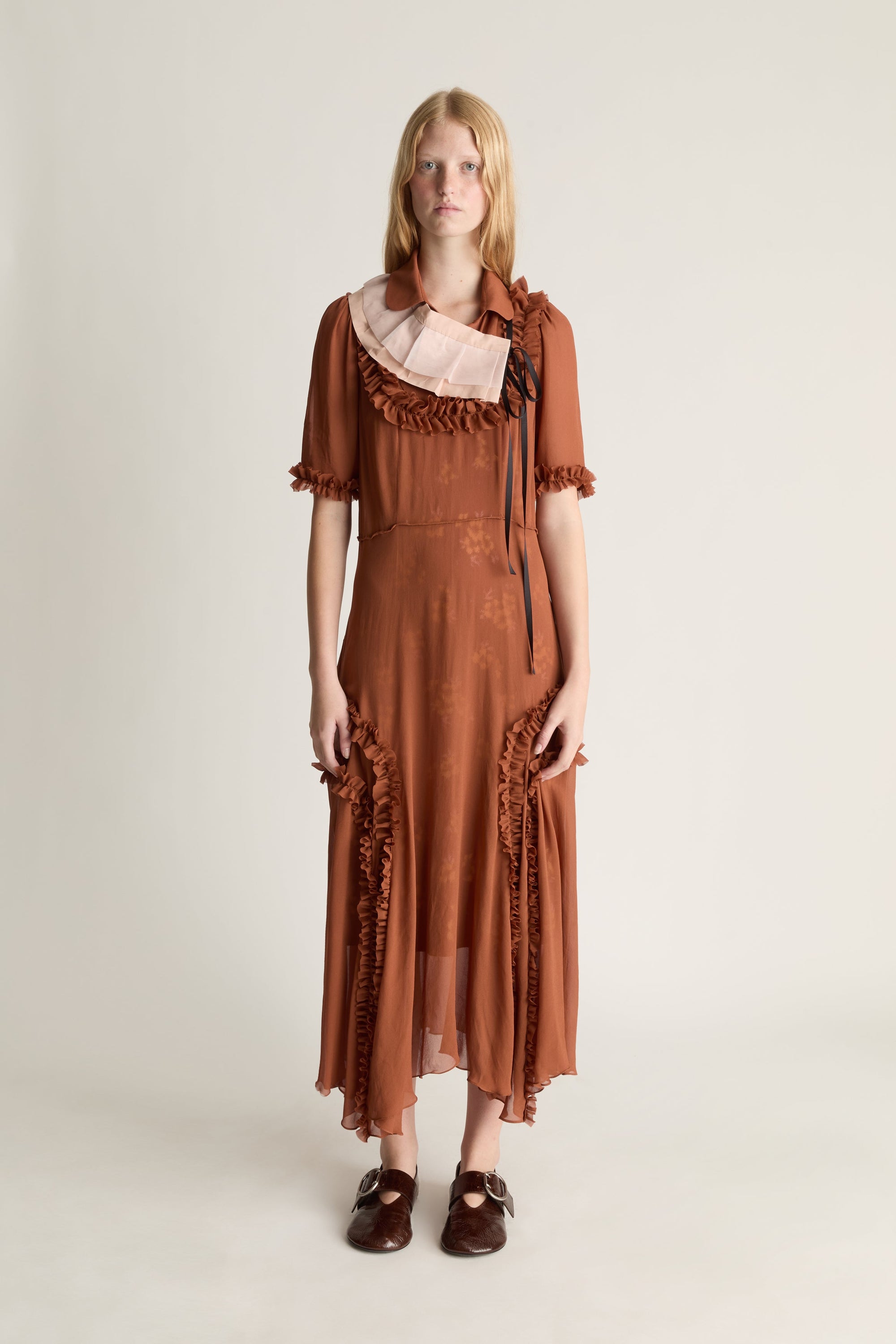Front view of a woman wearing a burnt orange georgette dress with frill details, shot against a studio background. 