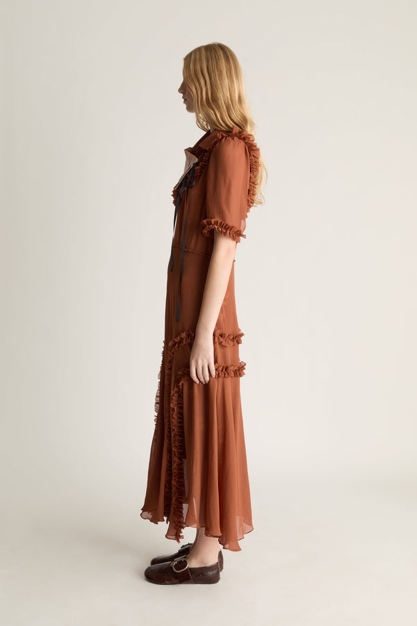 Profile view of a woman wearing a nutmeg coloured, georgette short sleeve dress and red shoes, shot against a studio background. 