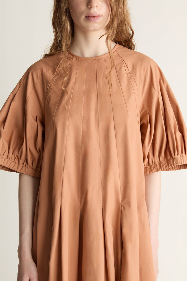 Close up image of a woman wearing a orange dress with gathered sleeves and panels down the front against a white studio background. 