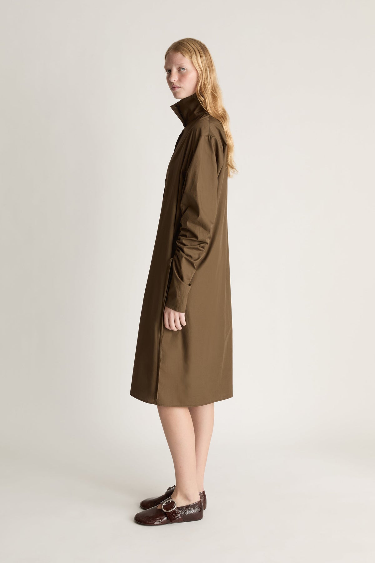 Woman standing profile wearing a long brown coat on a white background