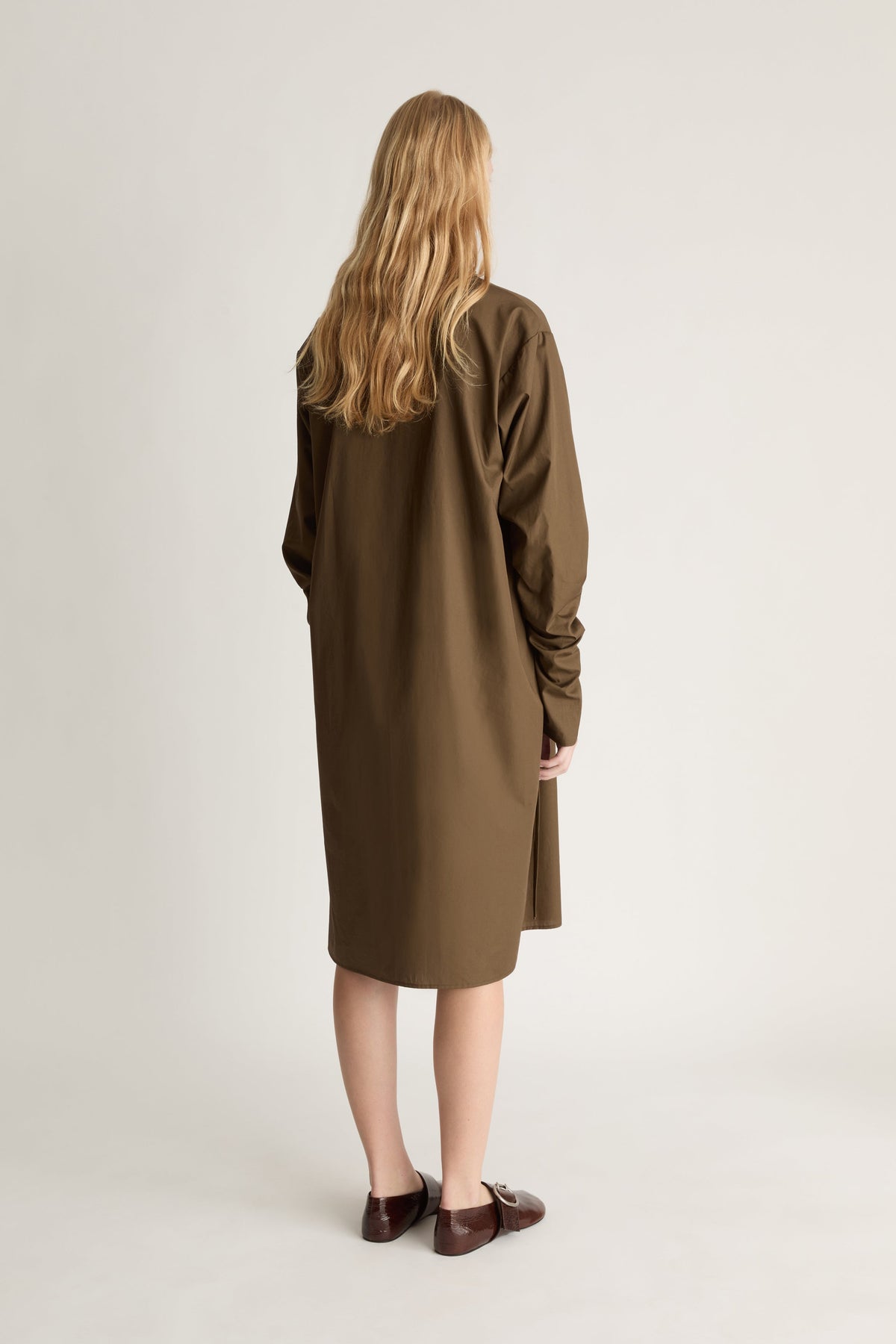 Back view of a person wearing a long brown coat standing against a plain background