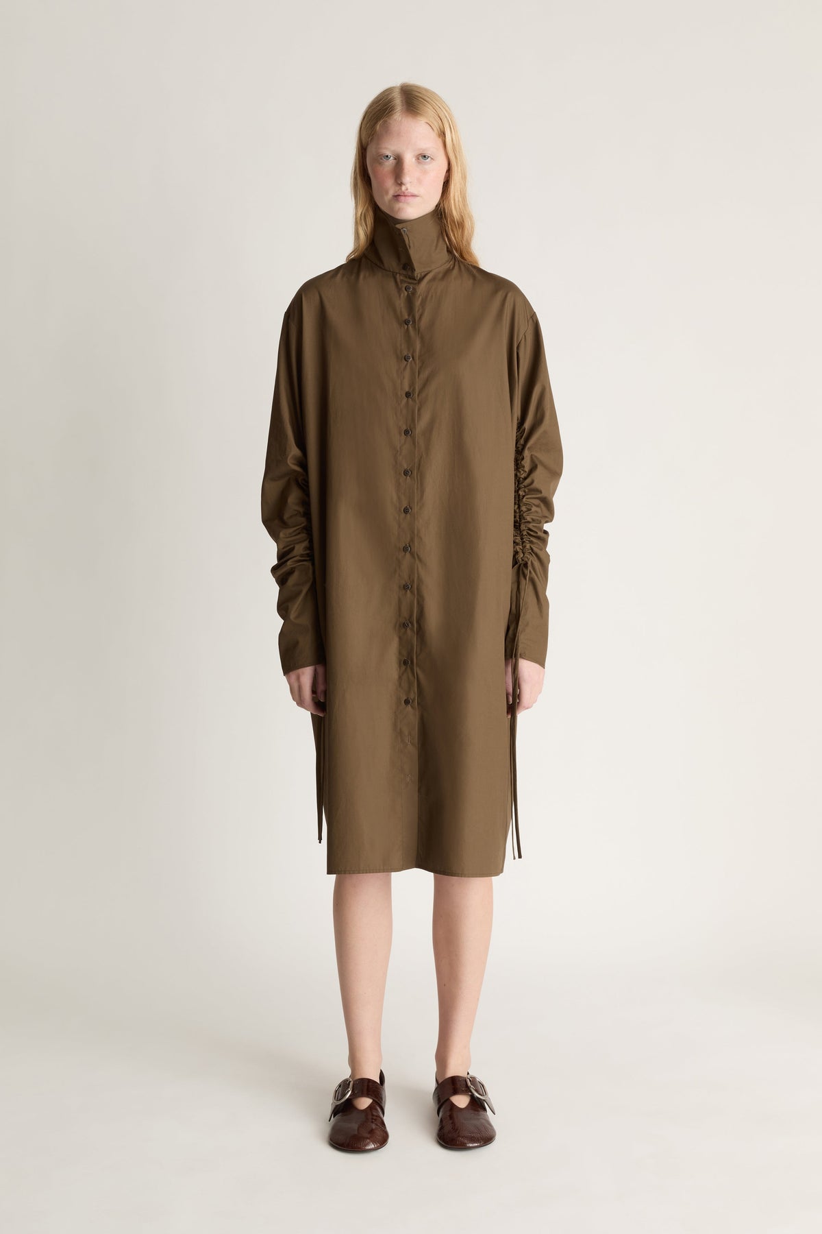 A model stands against a plain light beige studio background wearing a high-neck, long-sleeved brown dress with gathered sleeves and dark brown loafers.