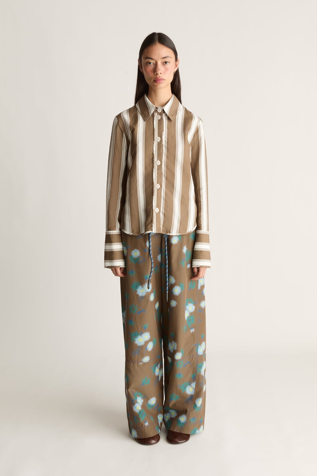 A model stands against a plain light gray studio background wearing a matching two-piece set consisting of a brown and beige vertically striped long-sleeve button-up shirt and wide-leg trousers with a brown base and light blue floral pattern.