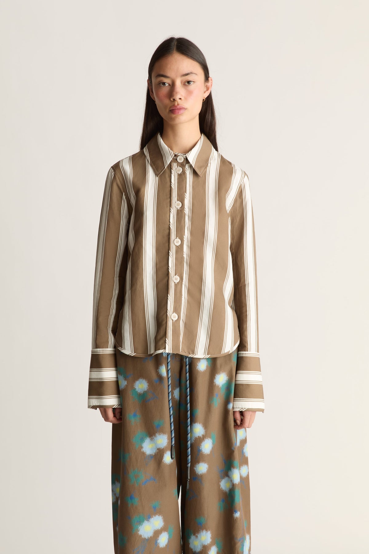 Close-up of a model against a plain light beige studio background wearing a brown and cream vertically striped button-up shirt, paired with brown wide-leg pants featuring a blue and white floral pattern.