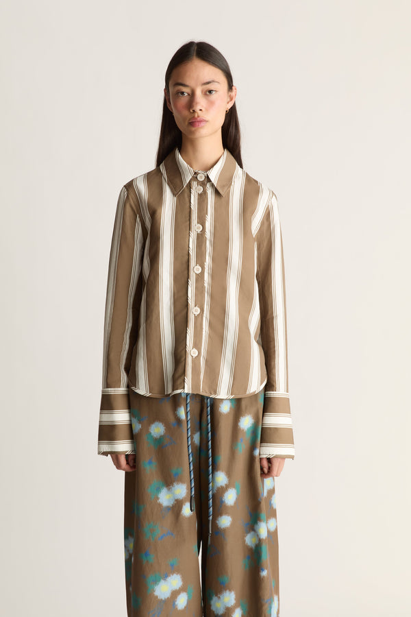 Close-up of a model against a plain light beige studio background wearing a brown and cream vertically striped button-up shirt, paired with brown wide-leg pants featuring a blue and white floral pattern.