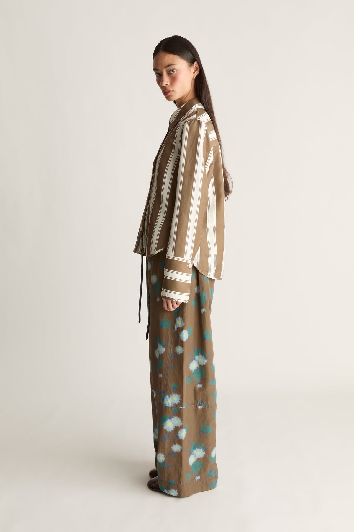Model wearing Tennant LS shirt— brown vertical-stripe shirt paired with brown floral-print wide-leg trousers, side profile