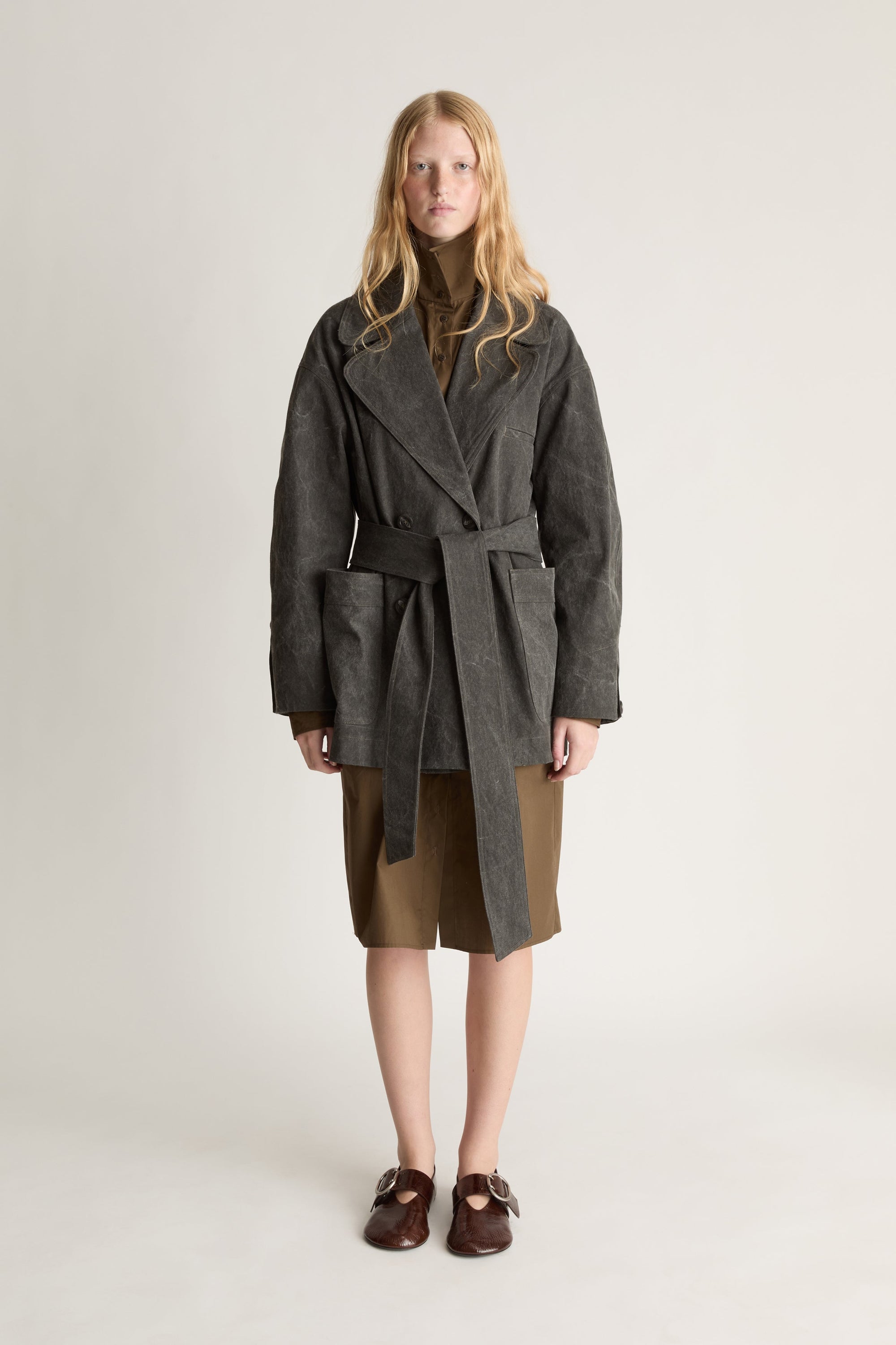 Front view of a woman wearing a charcoal pea coat over a khaki dress with red shoes, against a white studio background