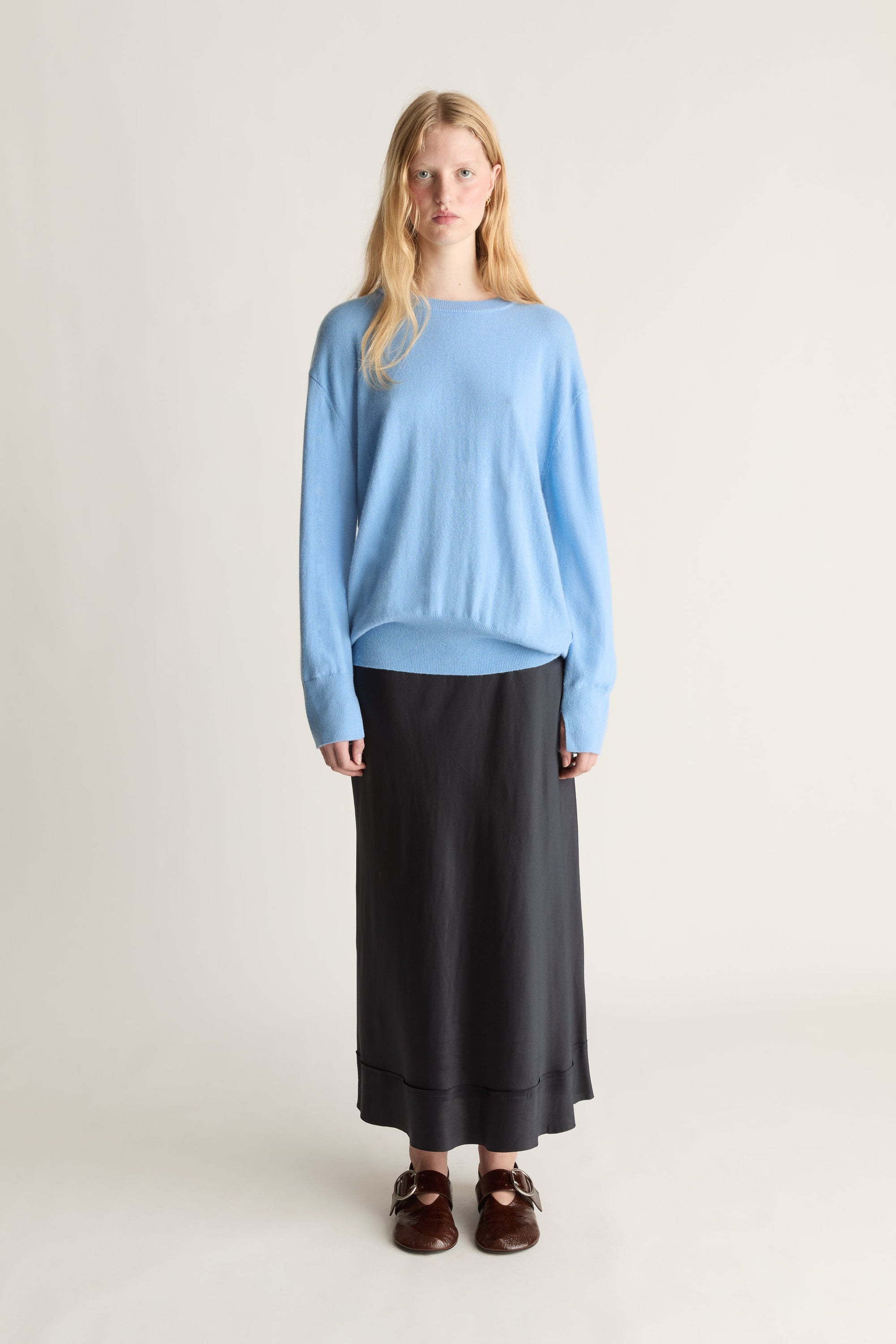 A woman stands against a plain light beige studio background wearing a light blue long-sleeve knit top, a long black skirt, and dark brown loafers.