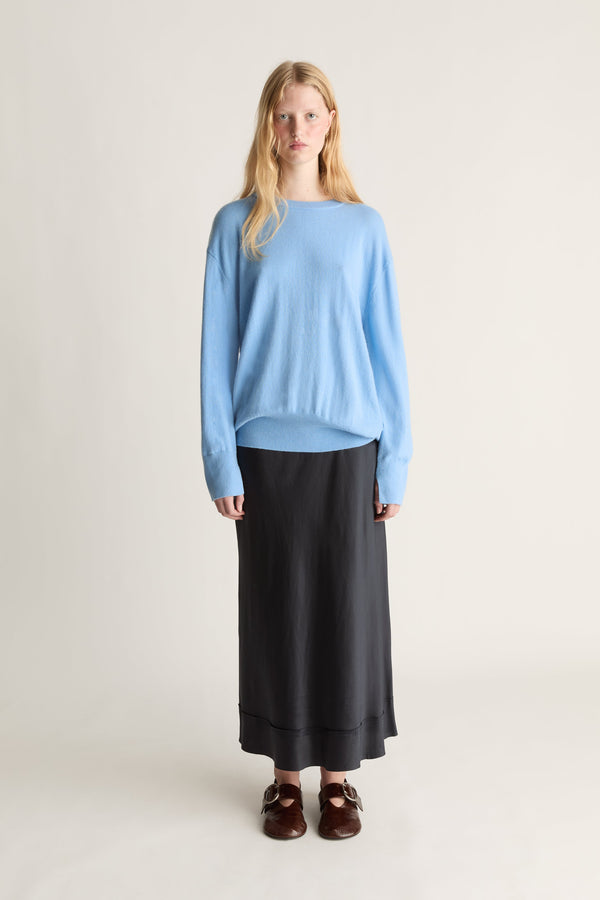 A woman stands against a plain light beige studio background wearing a light blue long-sleeve knit top, a long black skirt, and dark brown loafers.