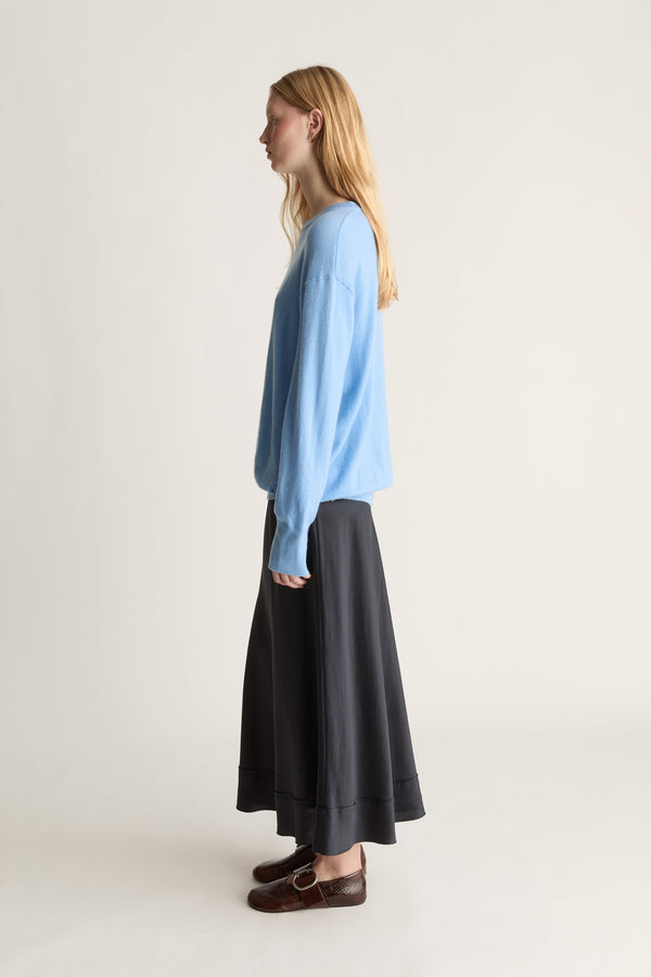 A woman stands in profile wearing a light blue long-sleeve knit top, a navy silk skirt, and brown leather loafers against a plain off-white studio background.