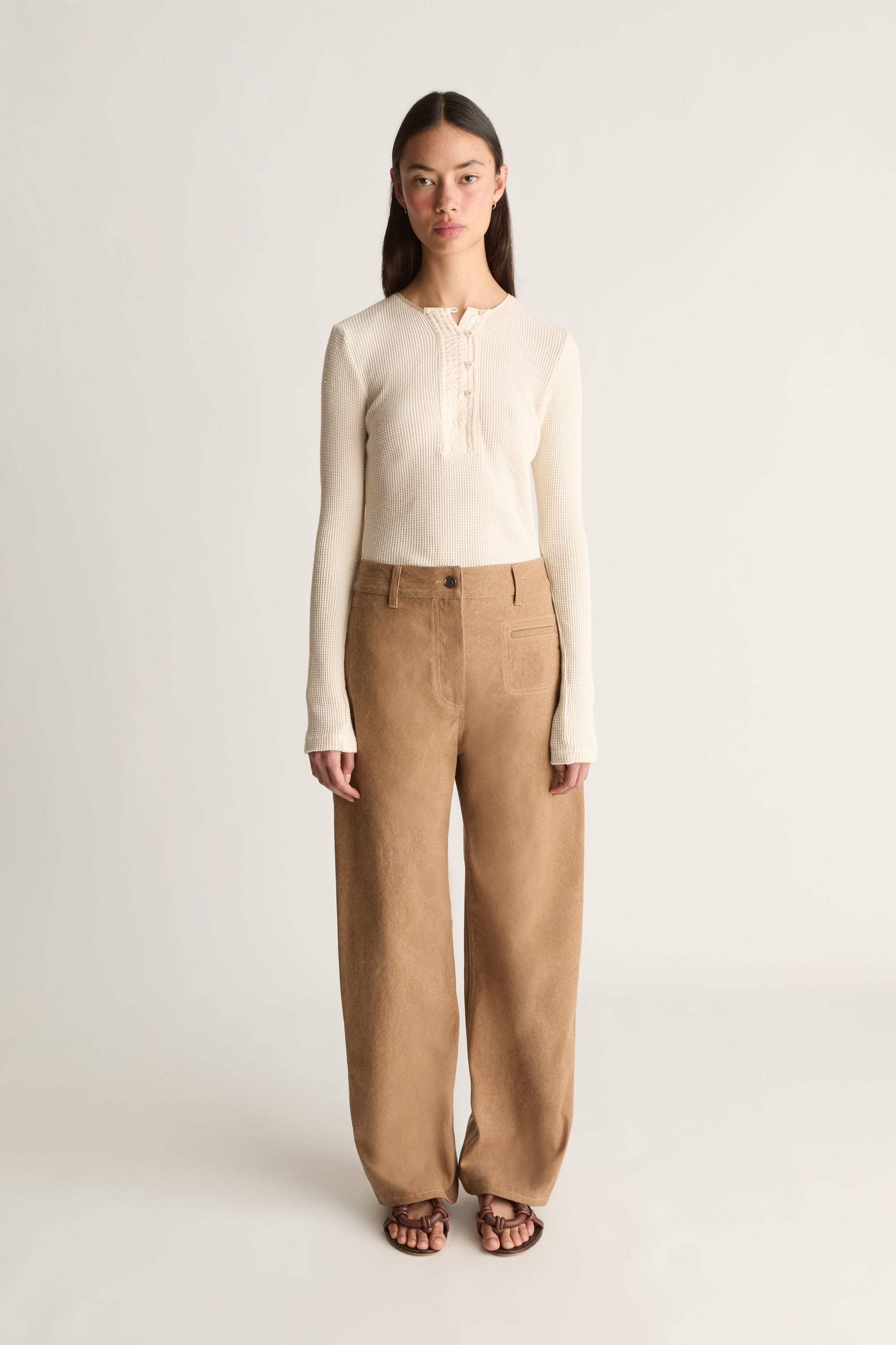 Front view of a model standing against a plain light gray studio background wearing a cream ribbed long-sleeve henley top and wide-leg tan trousers, with dark sandals.