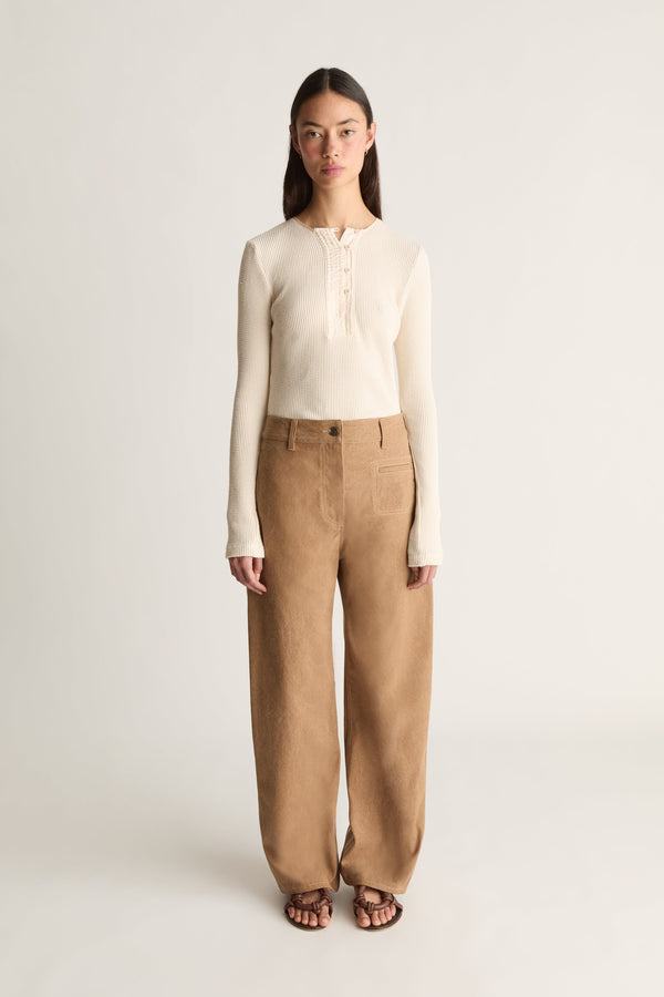 Front view of a model standing against a plain light gray studio background wearing a cream ribbed long-sleeve henley top and wide-leg tan trousers, with dark sandals.