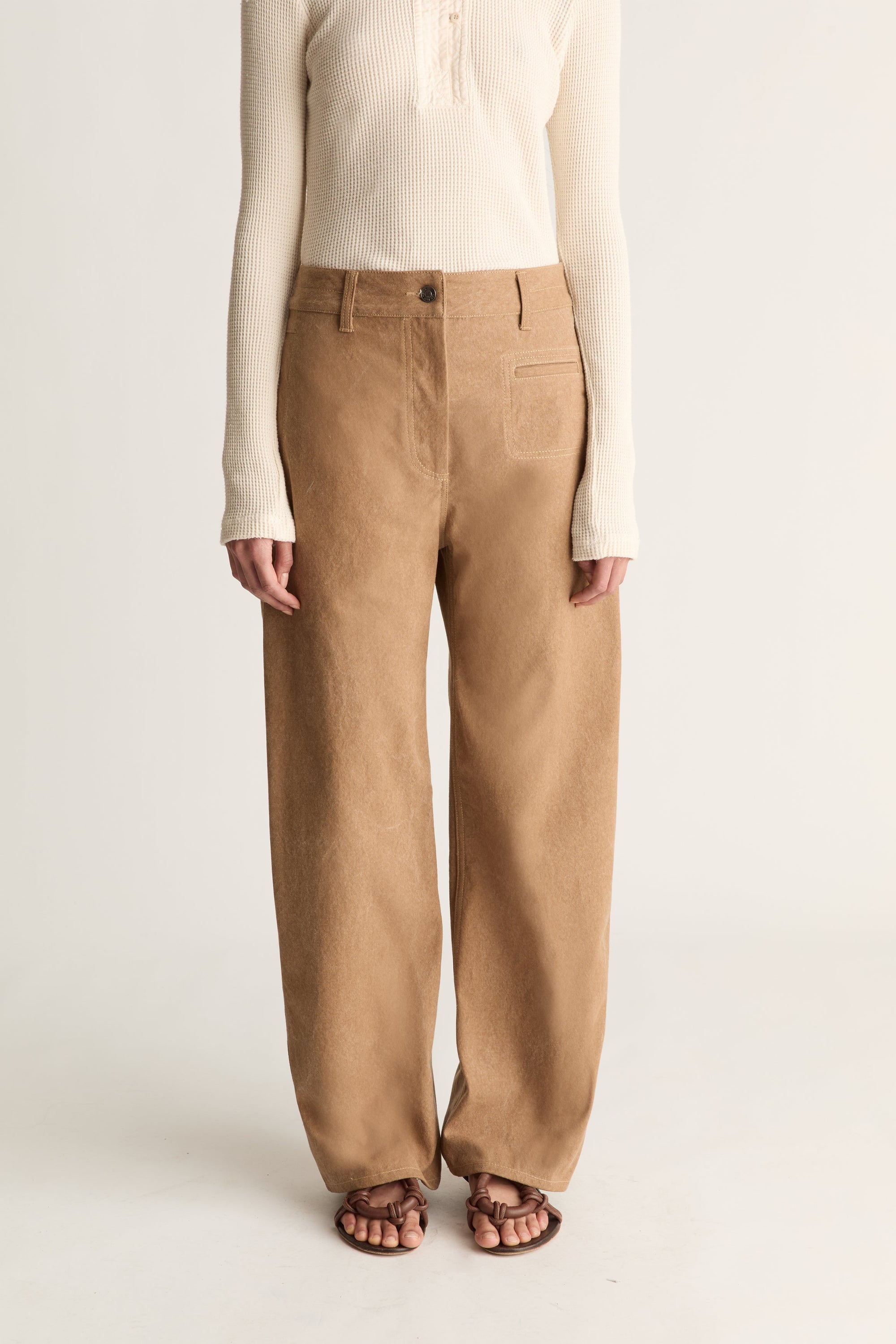 A model is shown from the chest down wearing a cream ribbed long-sleeve top and high-waisted, wide-leg tan corduroy trousers with brown sandals, set against a plain light gray studio background.