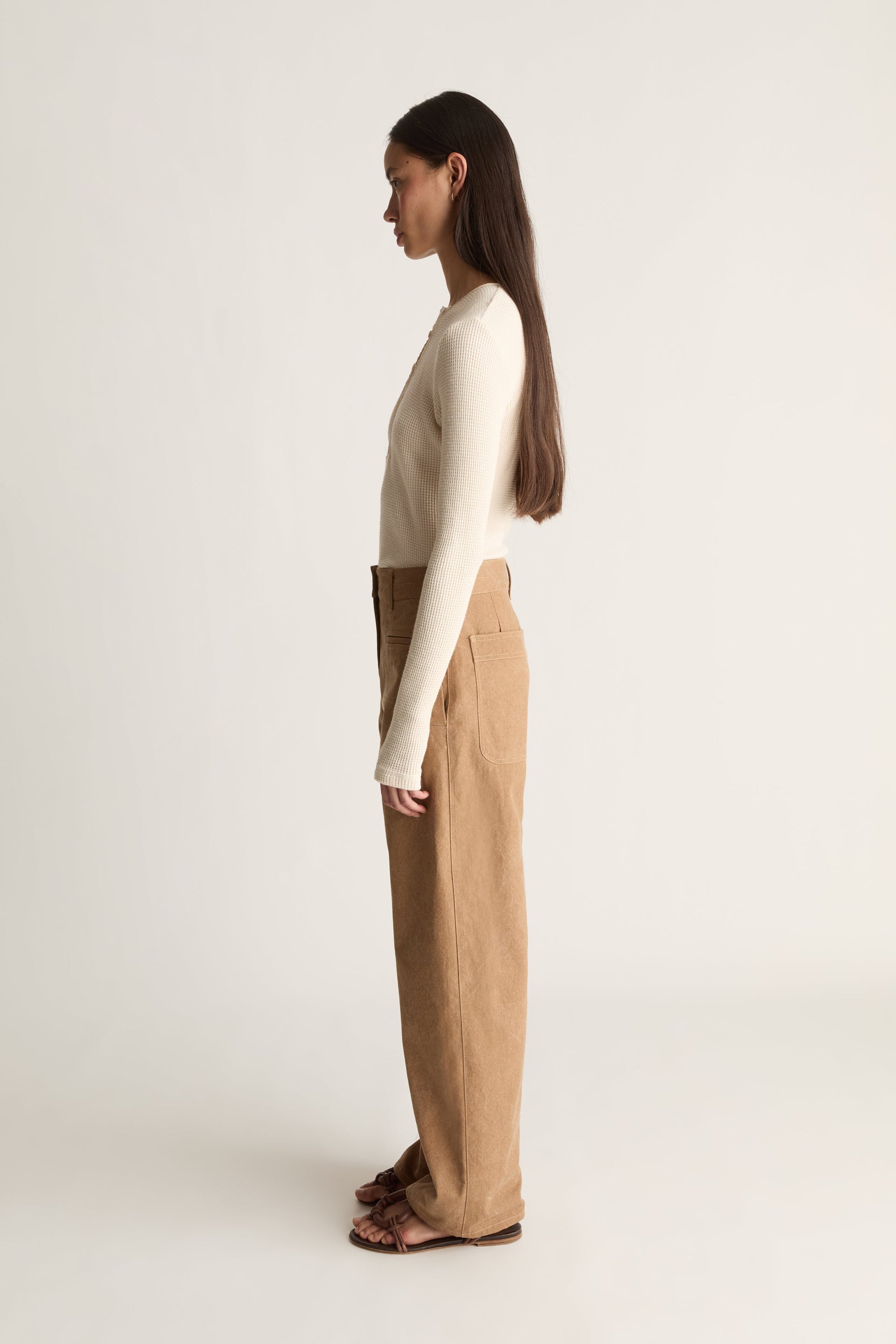 A woman stands in side profile wearing a fitted cream long-sleeve top and high-waisted tan wide-leg trousers, paired with dark brown sandals, against a plain light gray studio background.