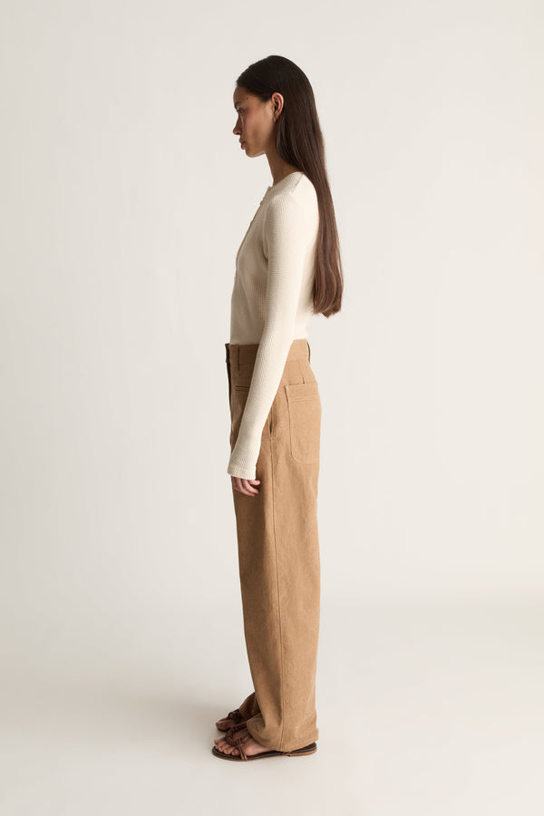A woman stands in side profile wearing a fitted cream long-sleeve top and high-waisted tan wide-leg trousers, paired with dark brown sandals, against a plain light gray studio background.