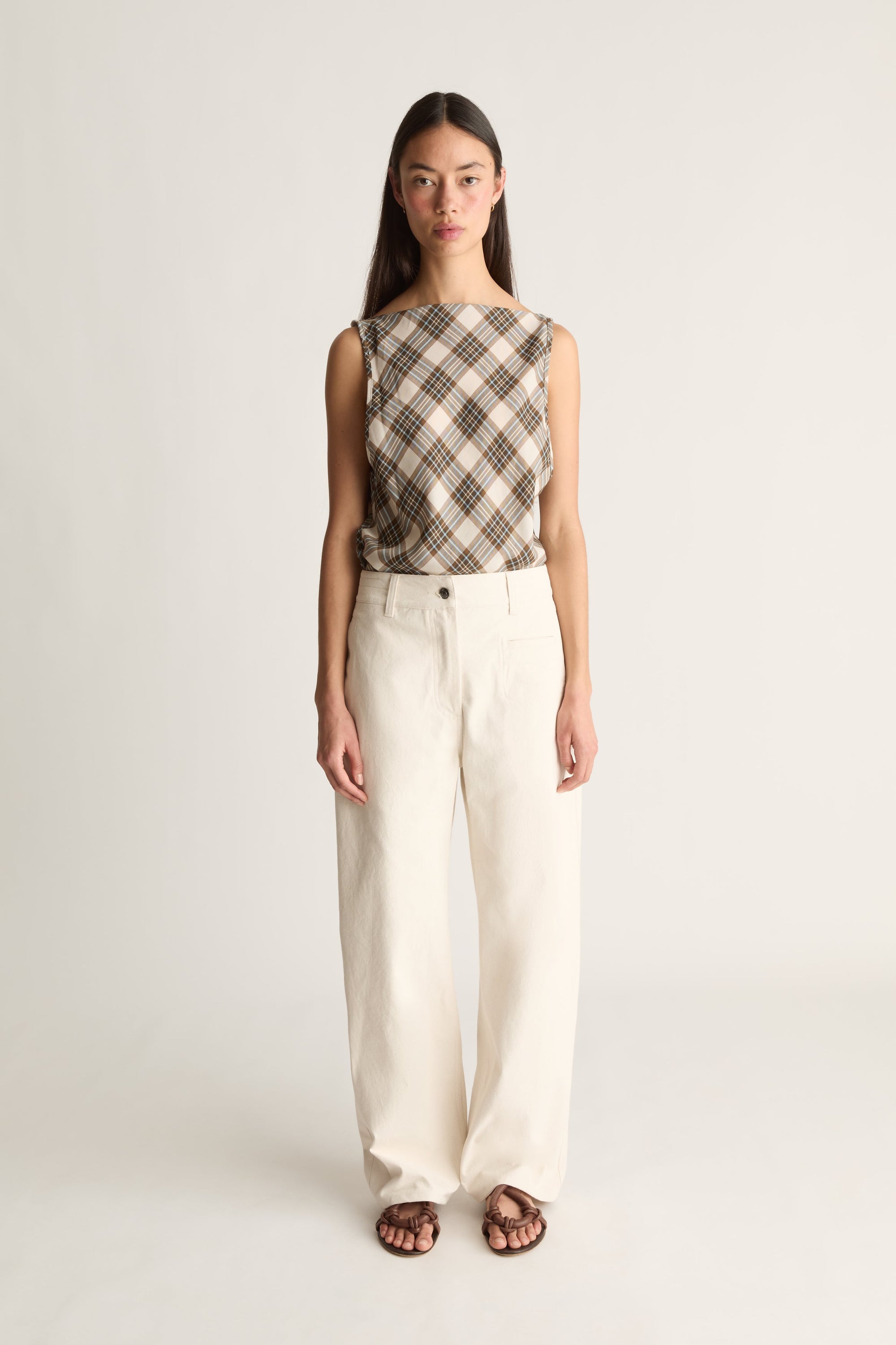 A model stands against a light gray studio background wearing a sleeveless plaid top in brown and beige with wide-leg cream trousers and dark sandals.