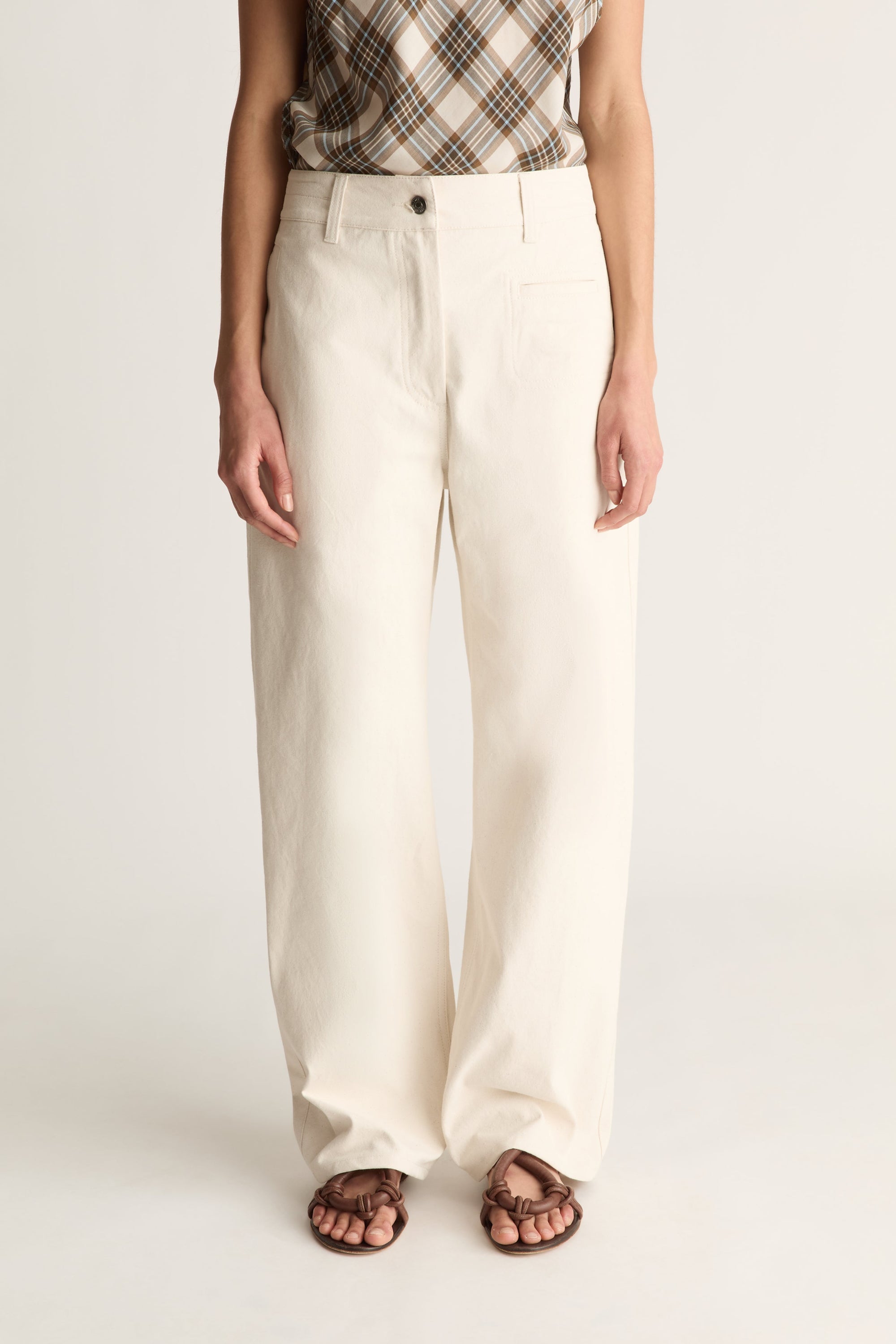 Close up of a model wearing a pair of off-white, wide-leg trousers with a brown and beige plaid top and brown sandals, shown from the waist down against a light gray studio background.