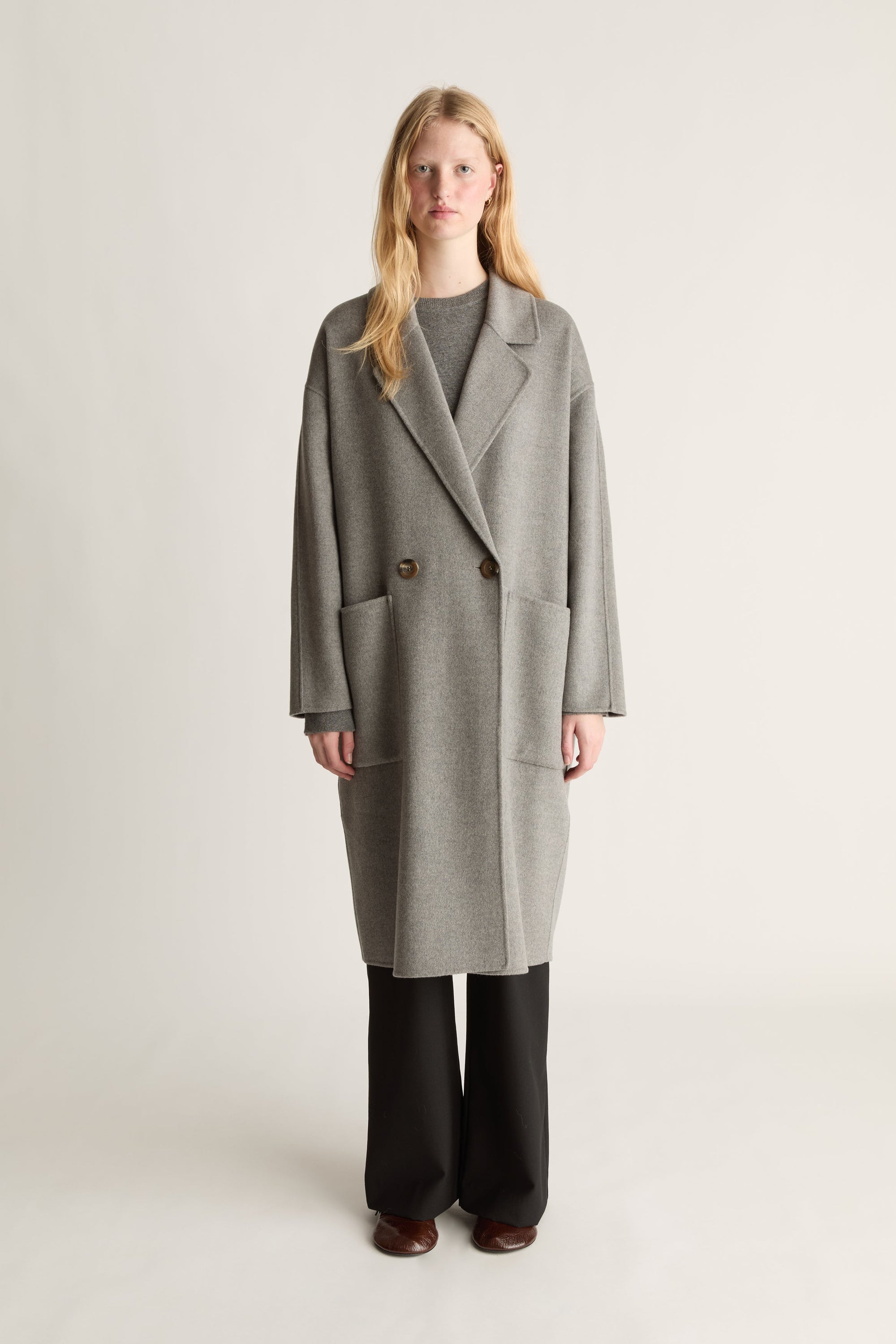 A woman stands against a plain light gray studio background wearing a long double-breasted gray coat over a dark top and black wide-leg pants.