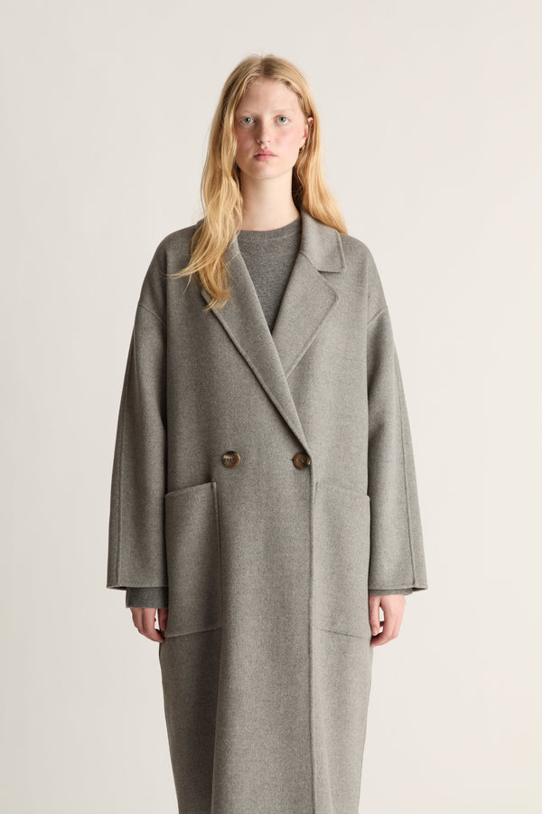 A close-up of a woman wearing a double-breasted gray wool-blend coat with wide lapels and dark buttons, photographed against a plain light gray studio background.