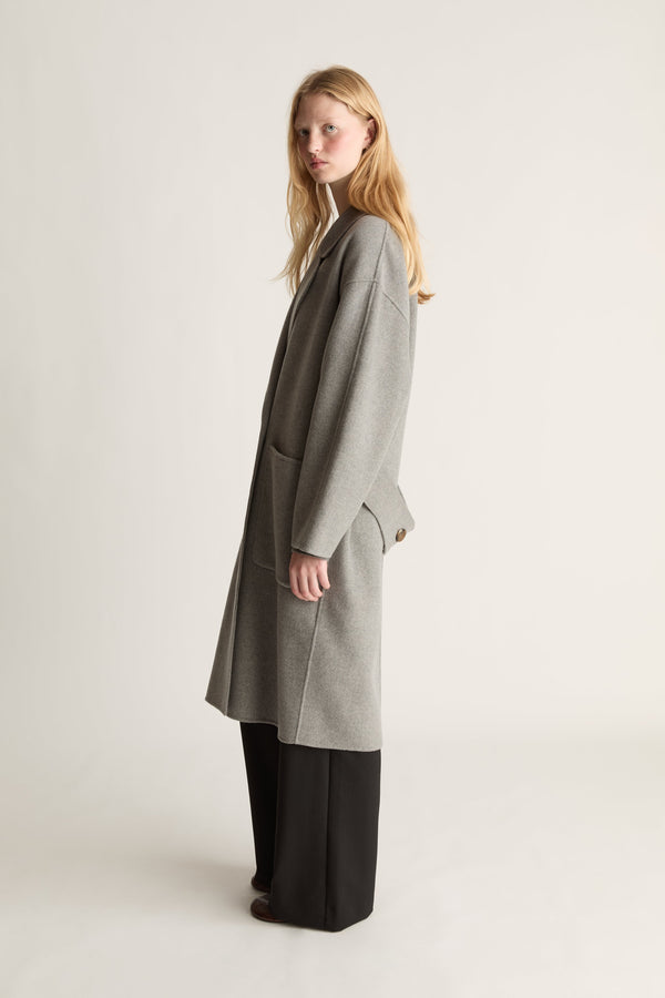 Side view of a woman wearing a  light gray wool-blend coat with black wide-leg trousers, standing in profile against a plain off-white studio background.
