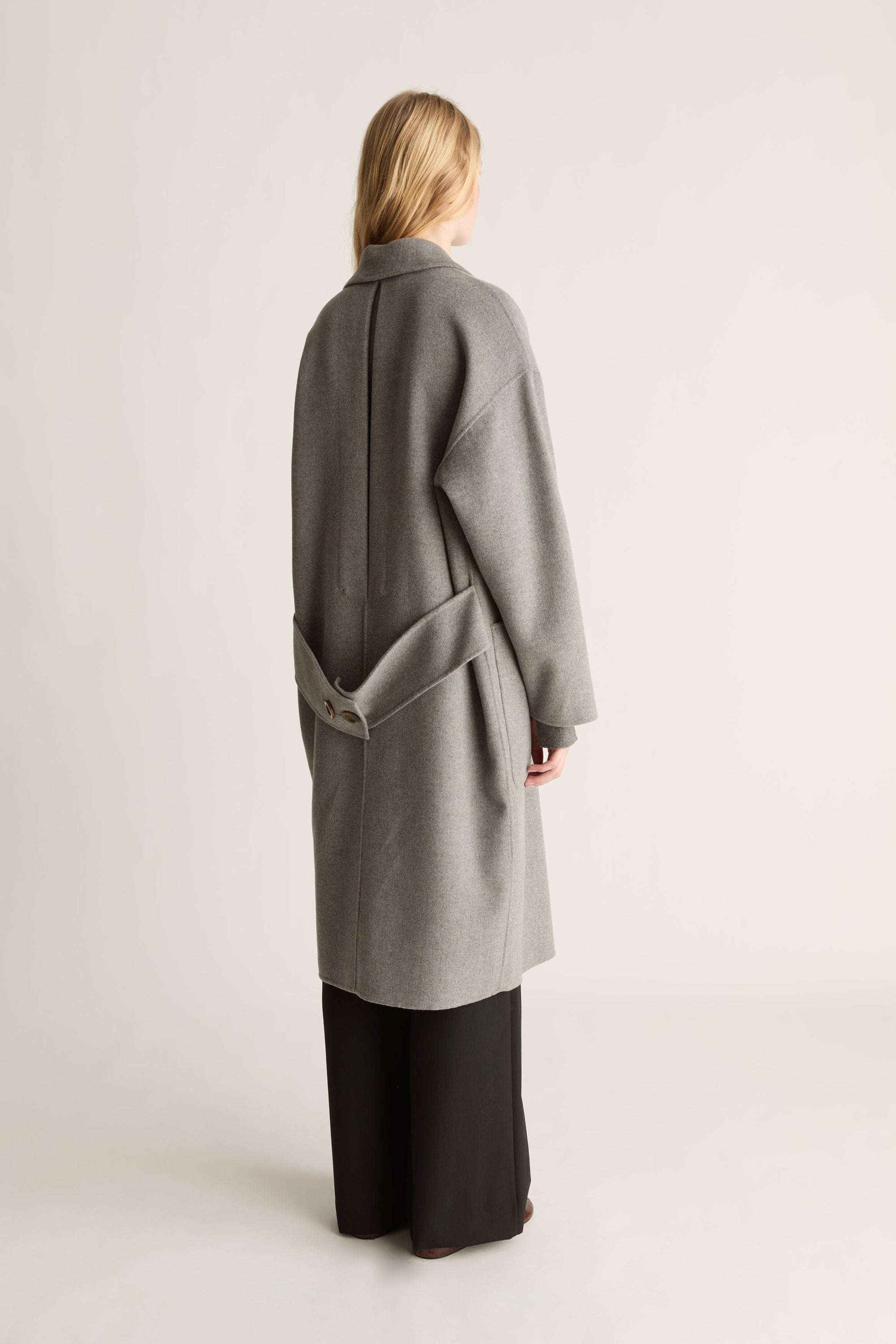 A woman shown from the back wearing a long, oversized light gray coat over black wide-leg pants, photographed against a plain light gray studio background.
