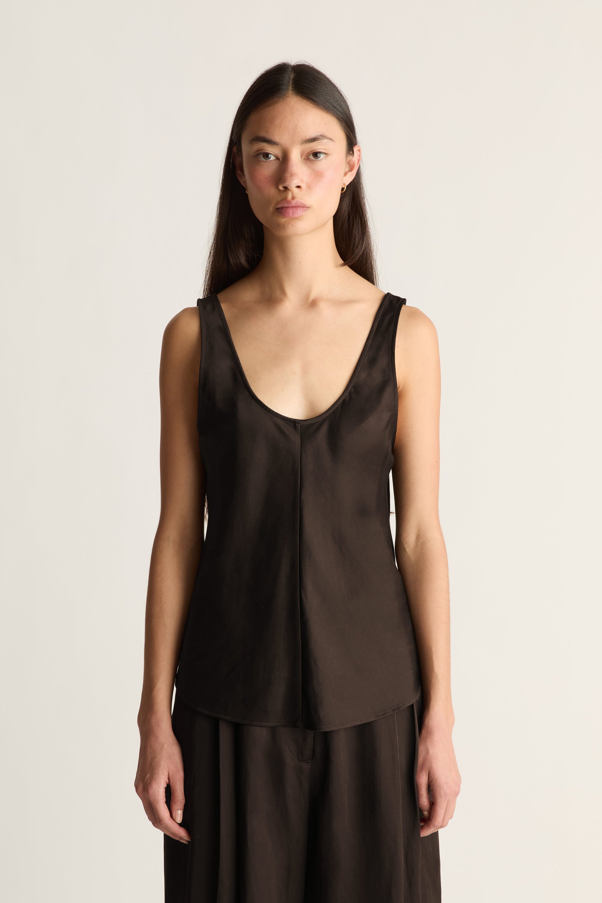 A model wearing a dark brown satin sleeveless top with a deep scoop neckline and a matching dark brown skirt, photographed against a plain light beige studio background.