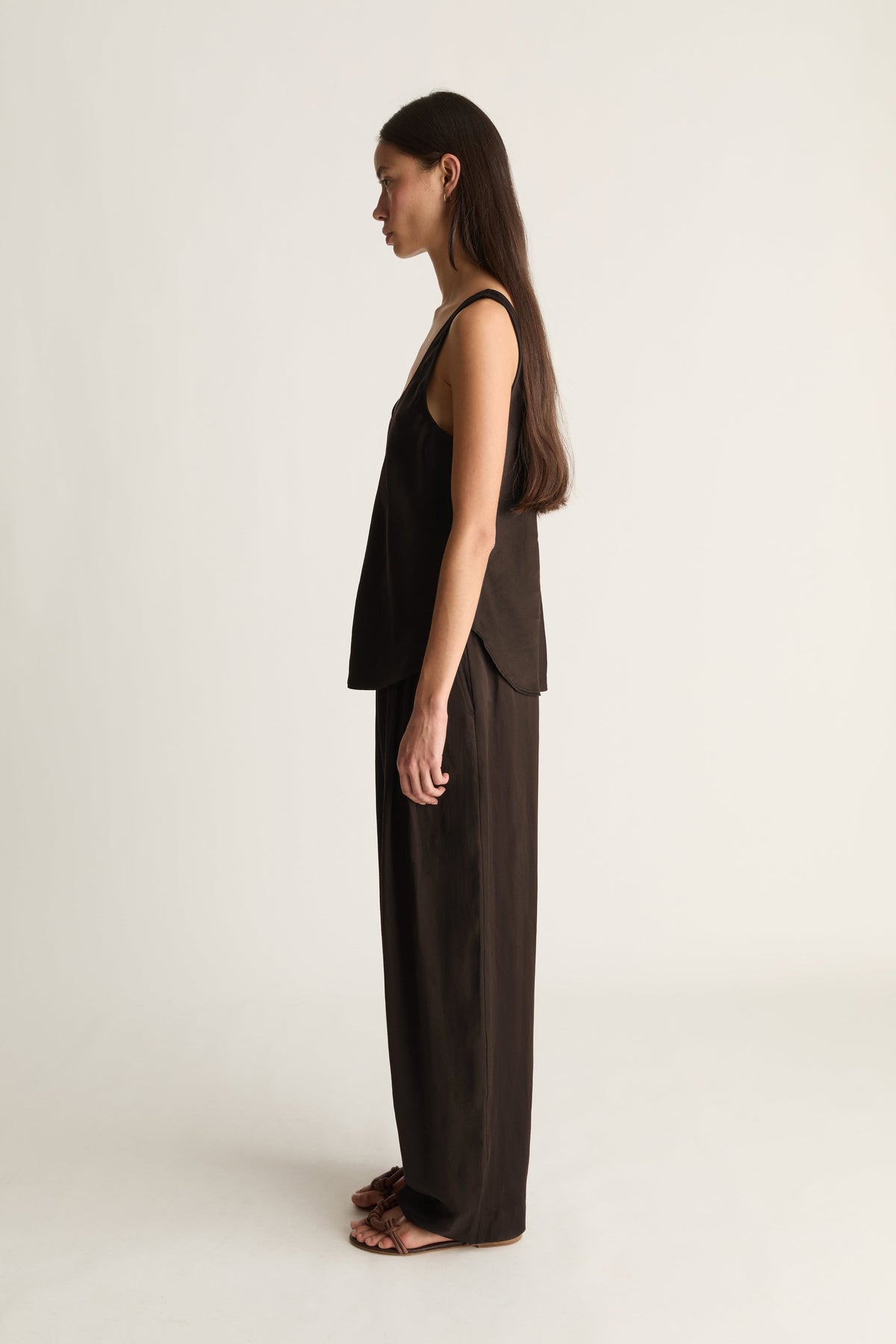 A woman stands in profile wearing a black sleeveless top and matching wide-leg pants against a plain light studio background.