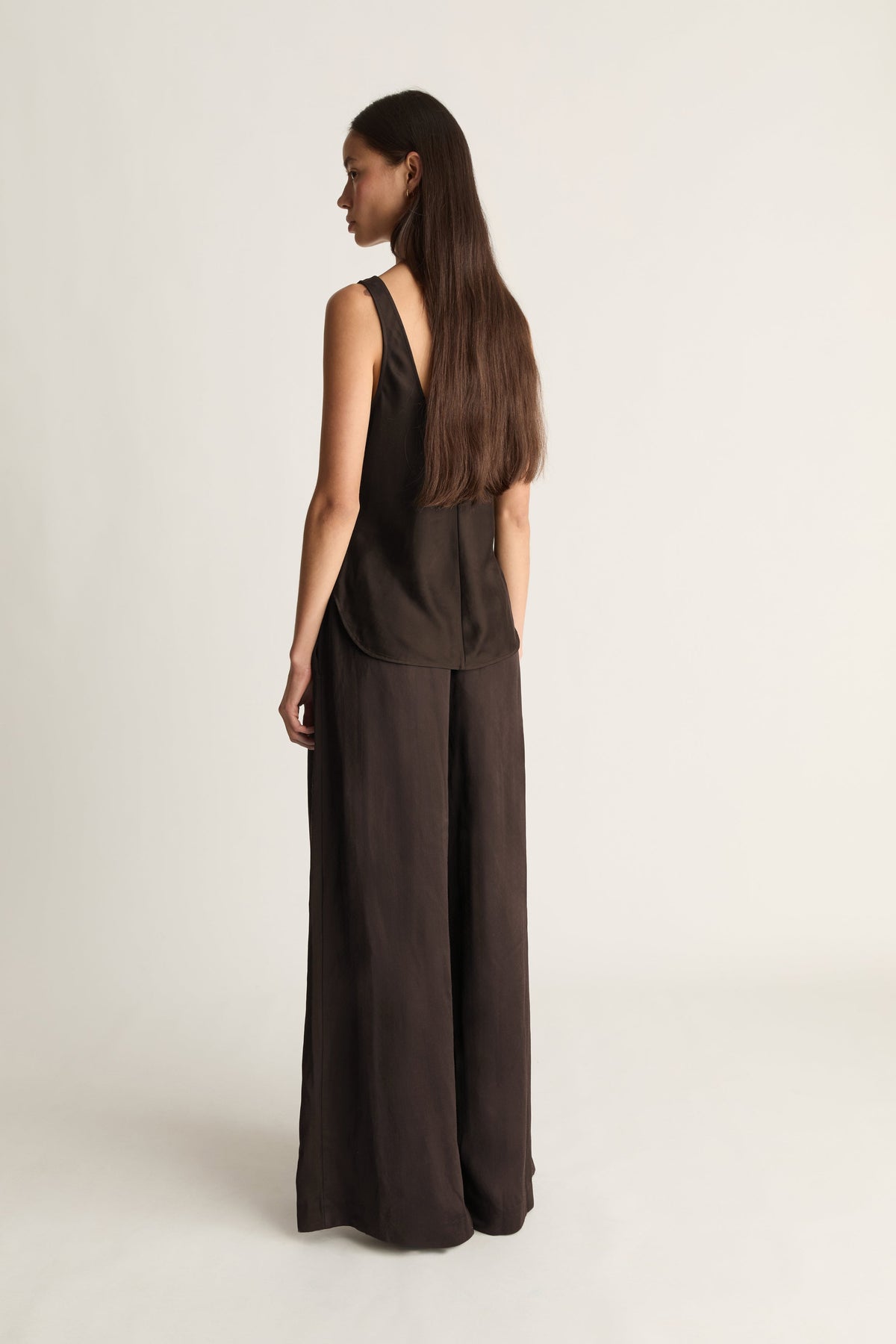 A woman with long dark brown hair is shown from the back wearing a dark brown sleeveless jumpsuit with a V-neckline and wide-leg pants, set against a plain light gray studio background.