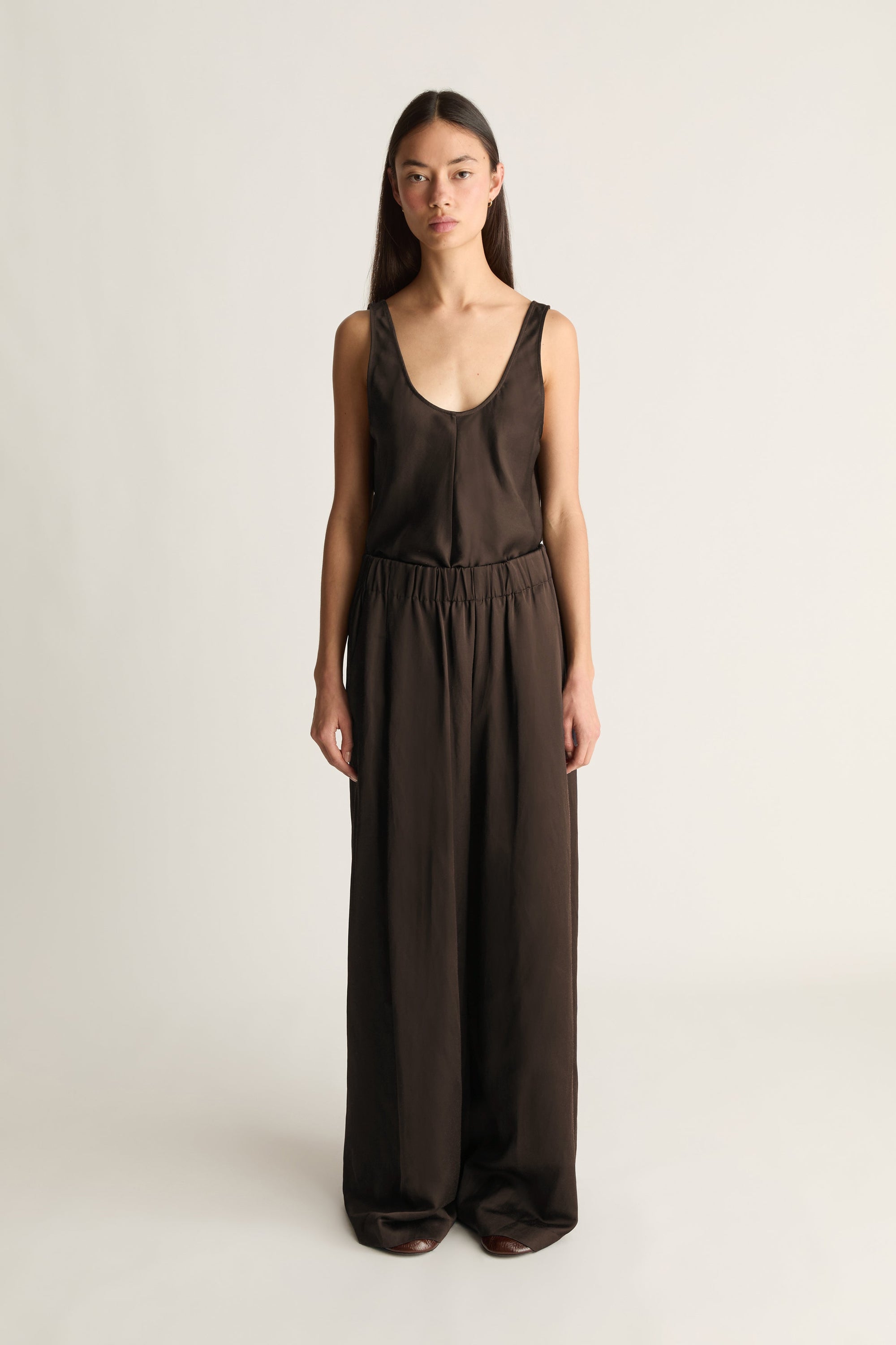 A woman modeling a dark brown sleeveless jumpsuit with a wide leg, deep scoop neckline, and elastic waistband, photographed against a plain light gray studio background.