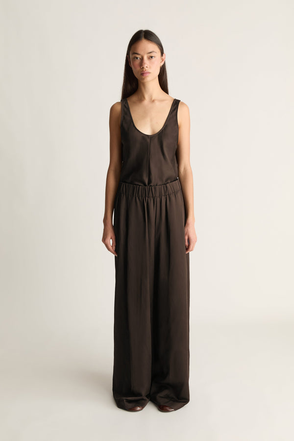 A woman modeling a dark brown sleeveless jumpsuit with a wide leg, deep scoop neckline, and elastic waistband, photographed against a plain light gray studio background.