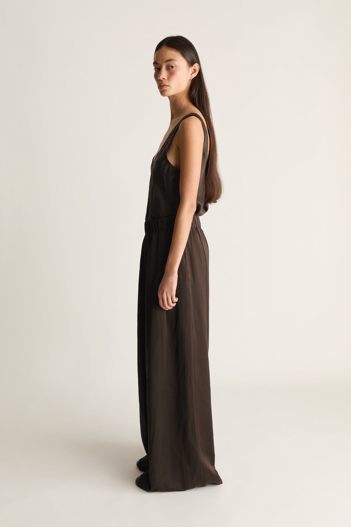 A woman stands in profile wearing a dark brown sleeveless maxi dress against a plain light beige studio background.