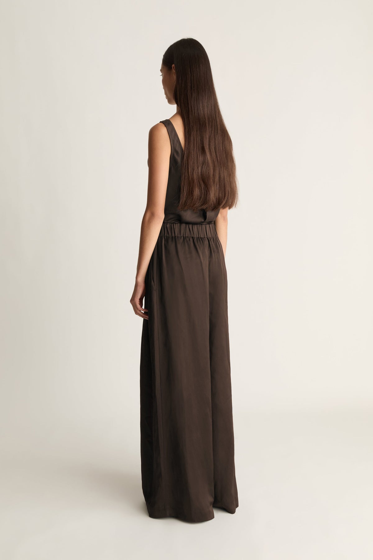 A woman with long dark brown hair is shown from the back wearing a sleeveless, wide-leg dark brown jumpsuit against a plain, light beige studio background.