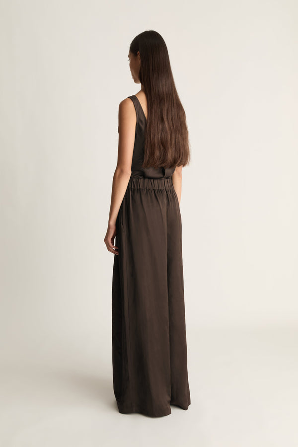 A woman with long dark brown hair is shown from the back wearing a sleeveless, wide-leg dark brown jumpsuit against a plain, light beige studio background.