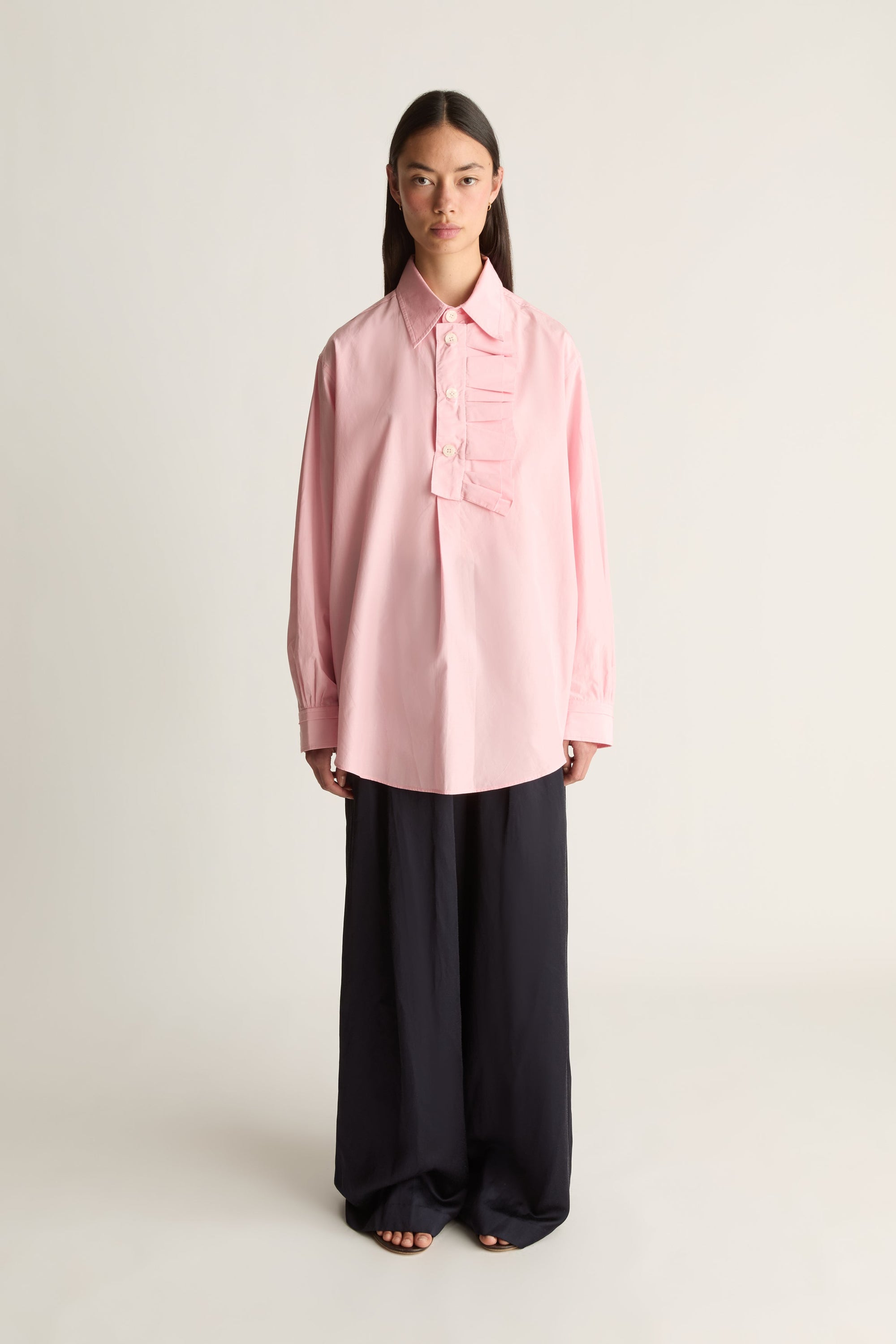 A model stands against a plain light beige studio background wearing a light pink silk long-sleeve button-up shirt with decorative ruffle details on the chest and dark wide-leg trousers.