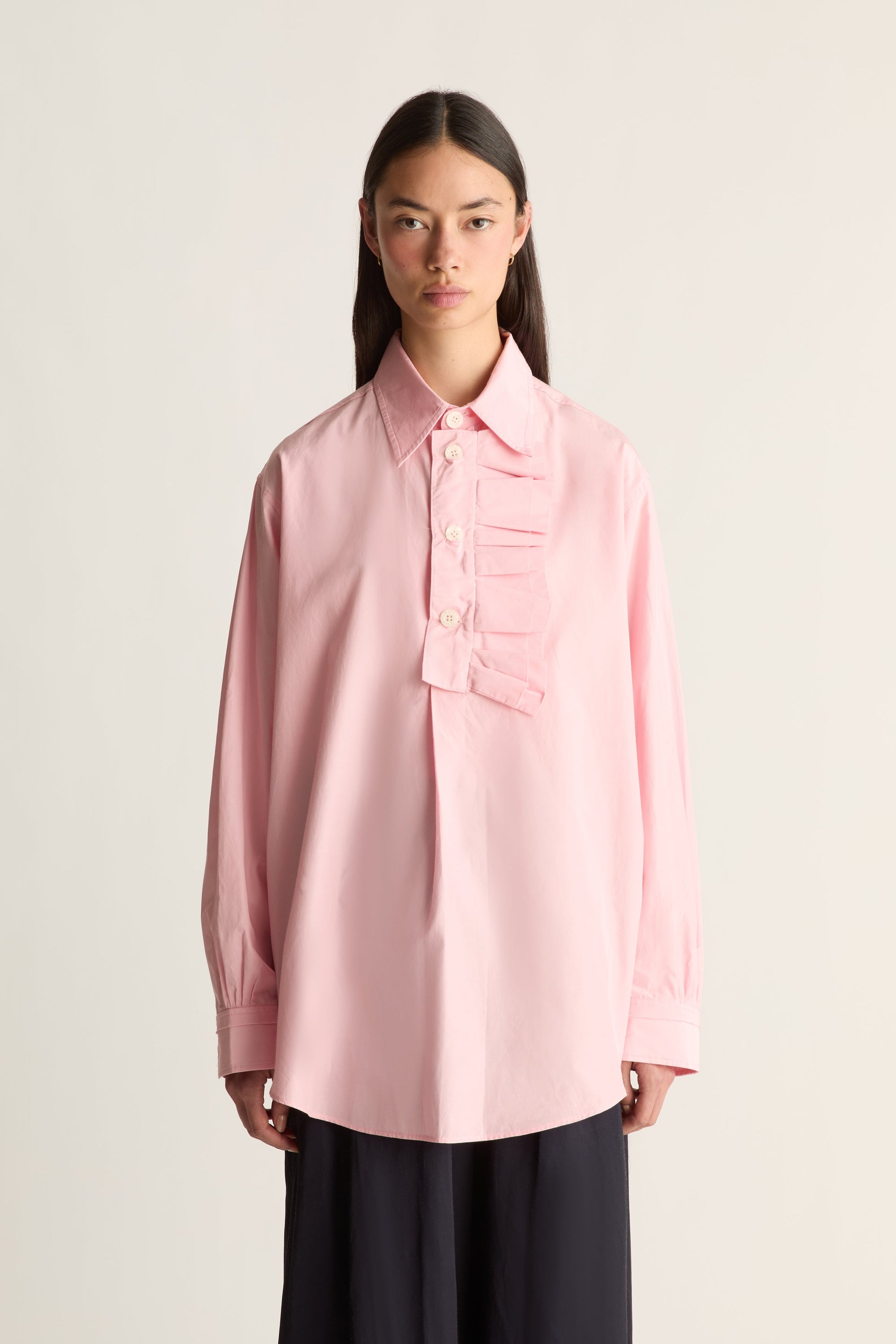Close up of a  model wearing a light pink long-sleeve button-up shirt with a decorative ruffle detail on the chest and dark wide-leg trousers, set against a plain light beige background.