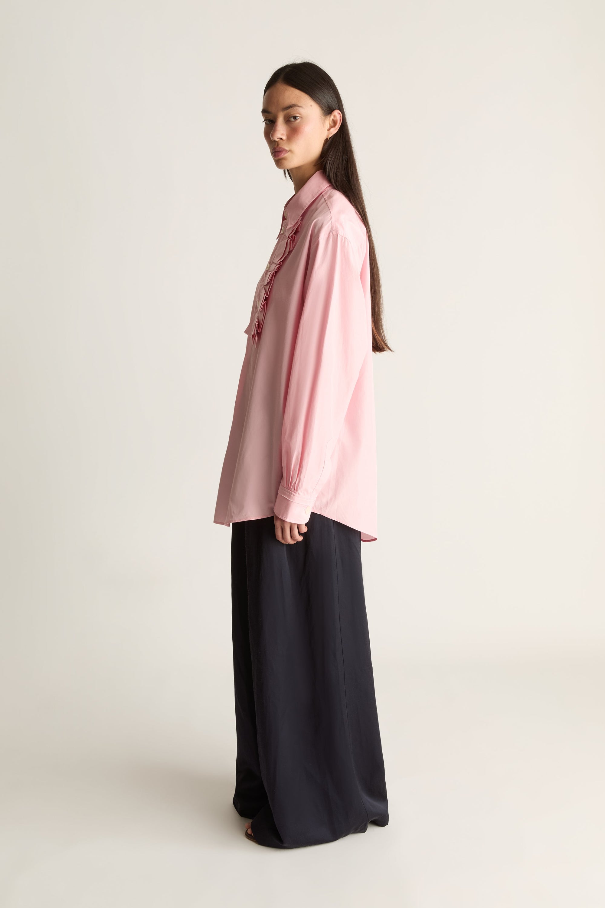 A woman stands in profile wearing a light pink long-sleeve blouse with a ruffled collar and dark wide-leg trousers against a plain light beige studio background.