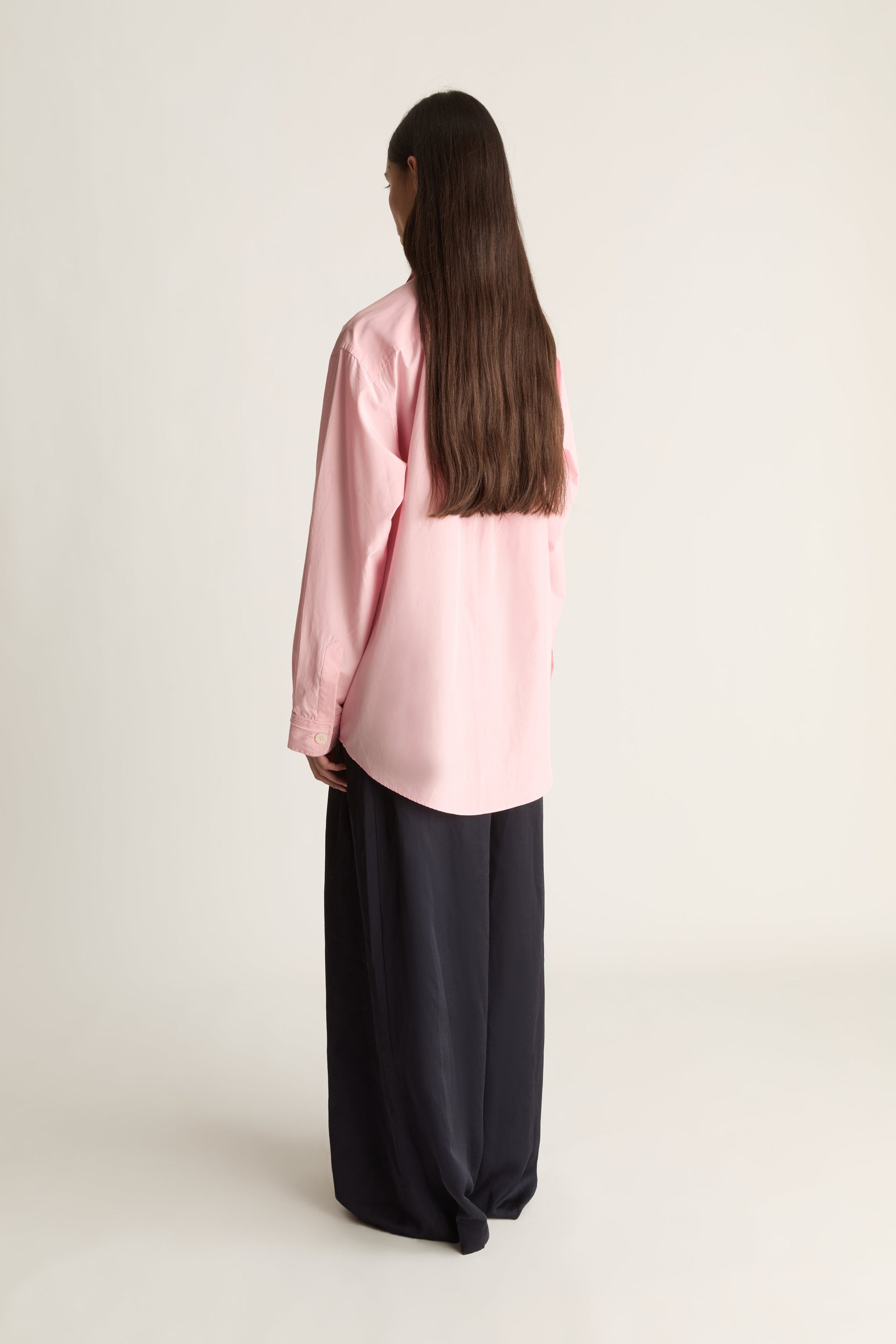 A woman with long dark brown hair is shown from behind wearing a light pink long-sleeve button-up shirt and wide-leg black trousers, standing against a plain off-white studio background.
