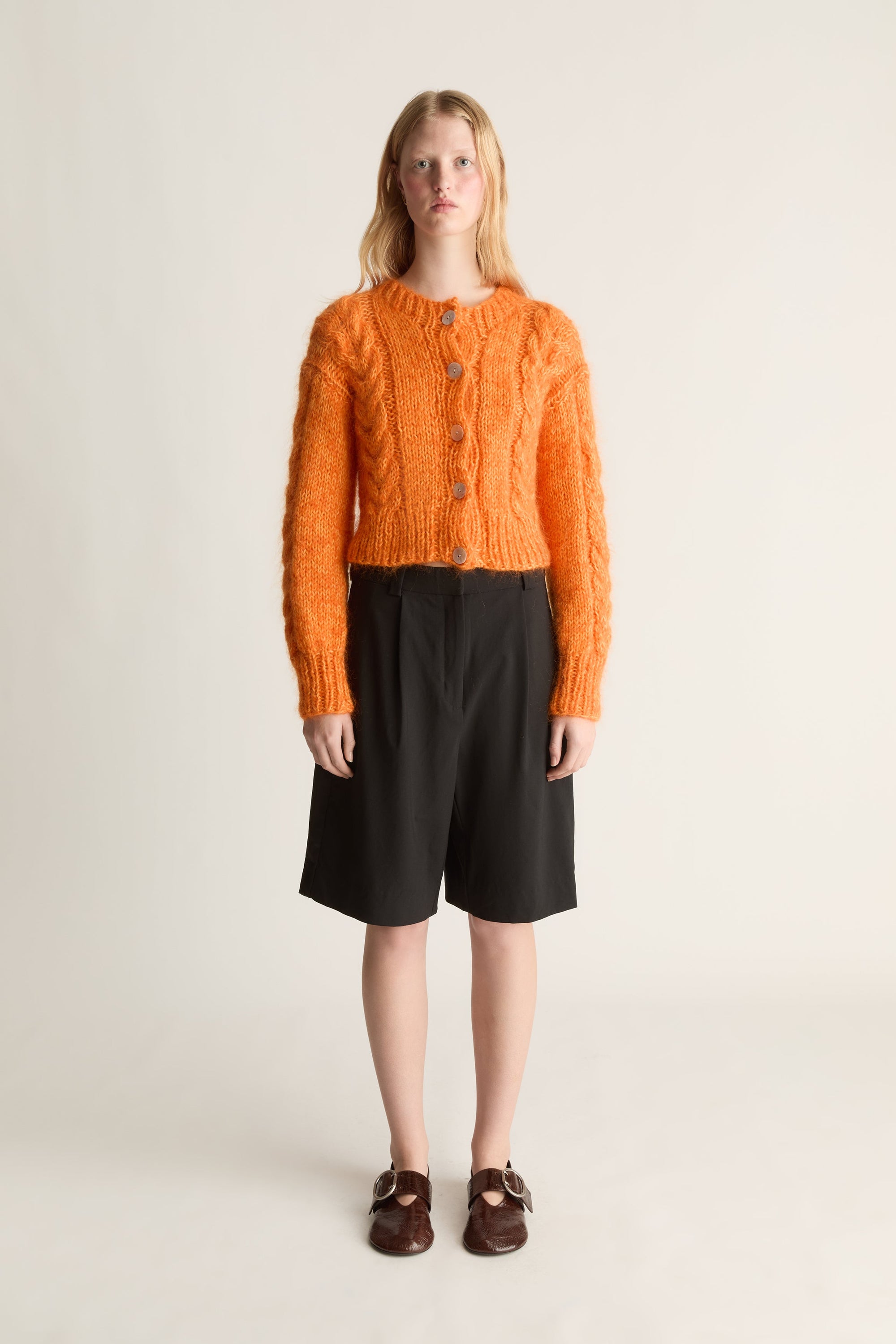 A model stands against a plain light beige studio background wearing a bright orange cable-knit cropped cardigan with buttons, black knee-length shorts, and dark brown loafers.