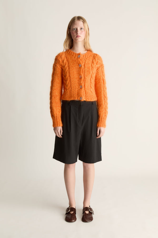 A model stands against a plain light beige studio background wearing a bright orange cable-knit cropped cardigan with buttons, black knee-length shorts, and dark brown loafers.