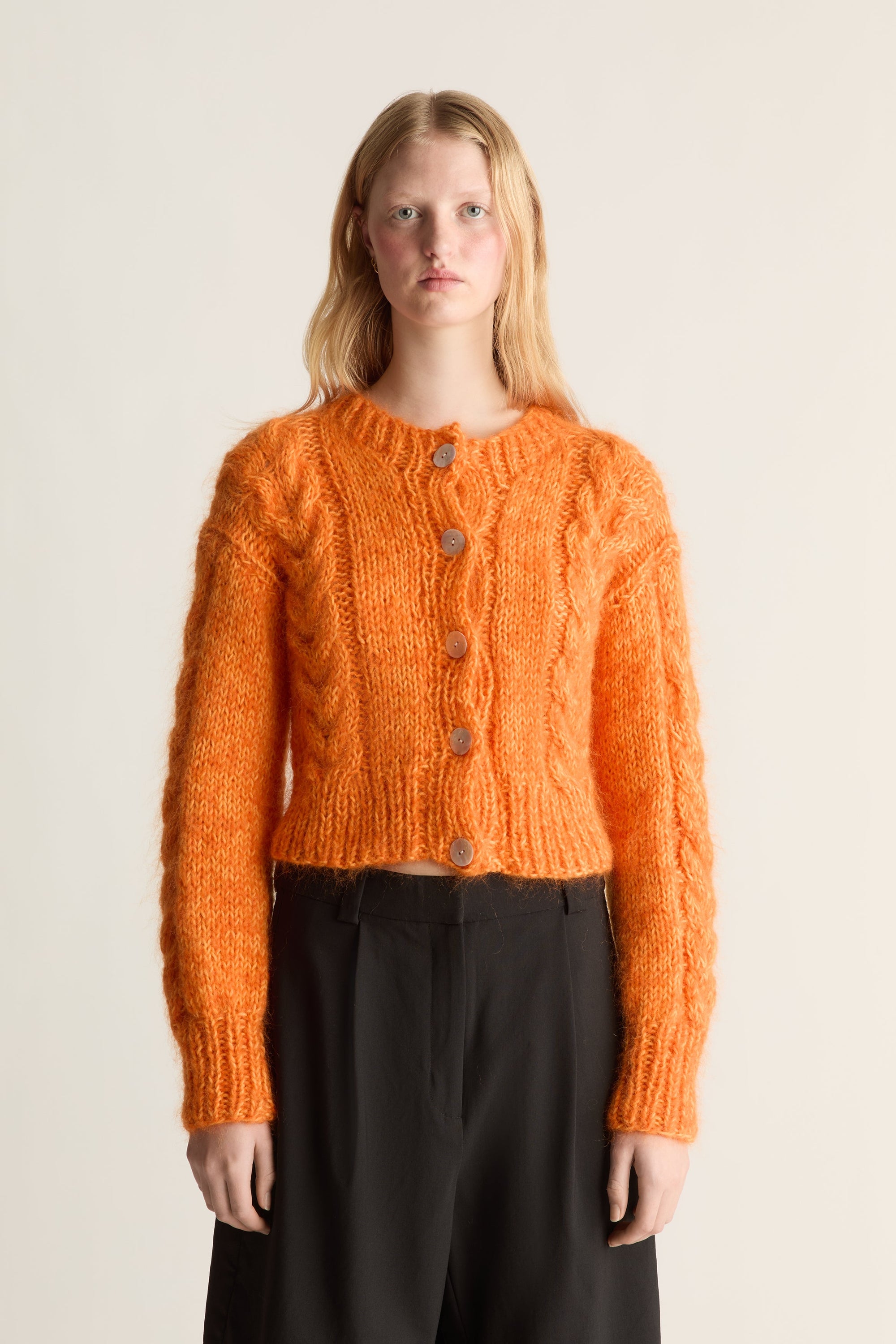 Close up of a woman modeling a cropped, cable-knit orange cardigan sweater with brown buttons, paired with black trousers, against a plain beige studio background.