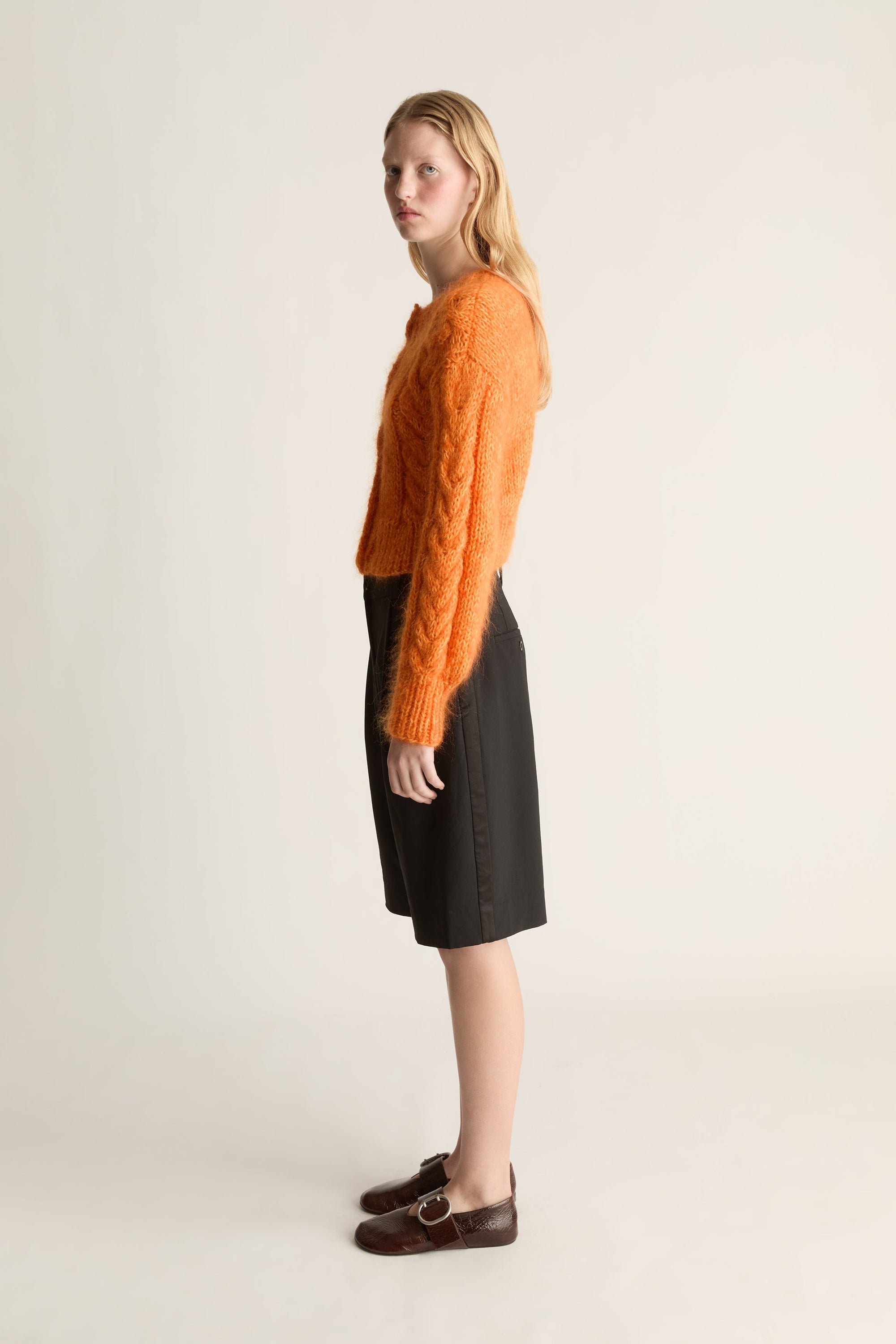 A woman stands in profile wearing a fuzzy orange cropped sweater, a black knee-length skirt, and dark brown loafers against a plain light beige studio background.