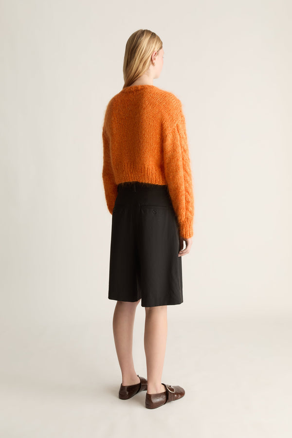 A woman is shown from behind wearing a fuzzy orange cropped sweater, black knee-length shorts, and brown leather loafers against a plain light beige studio background.