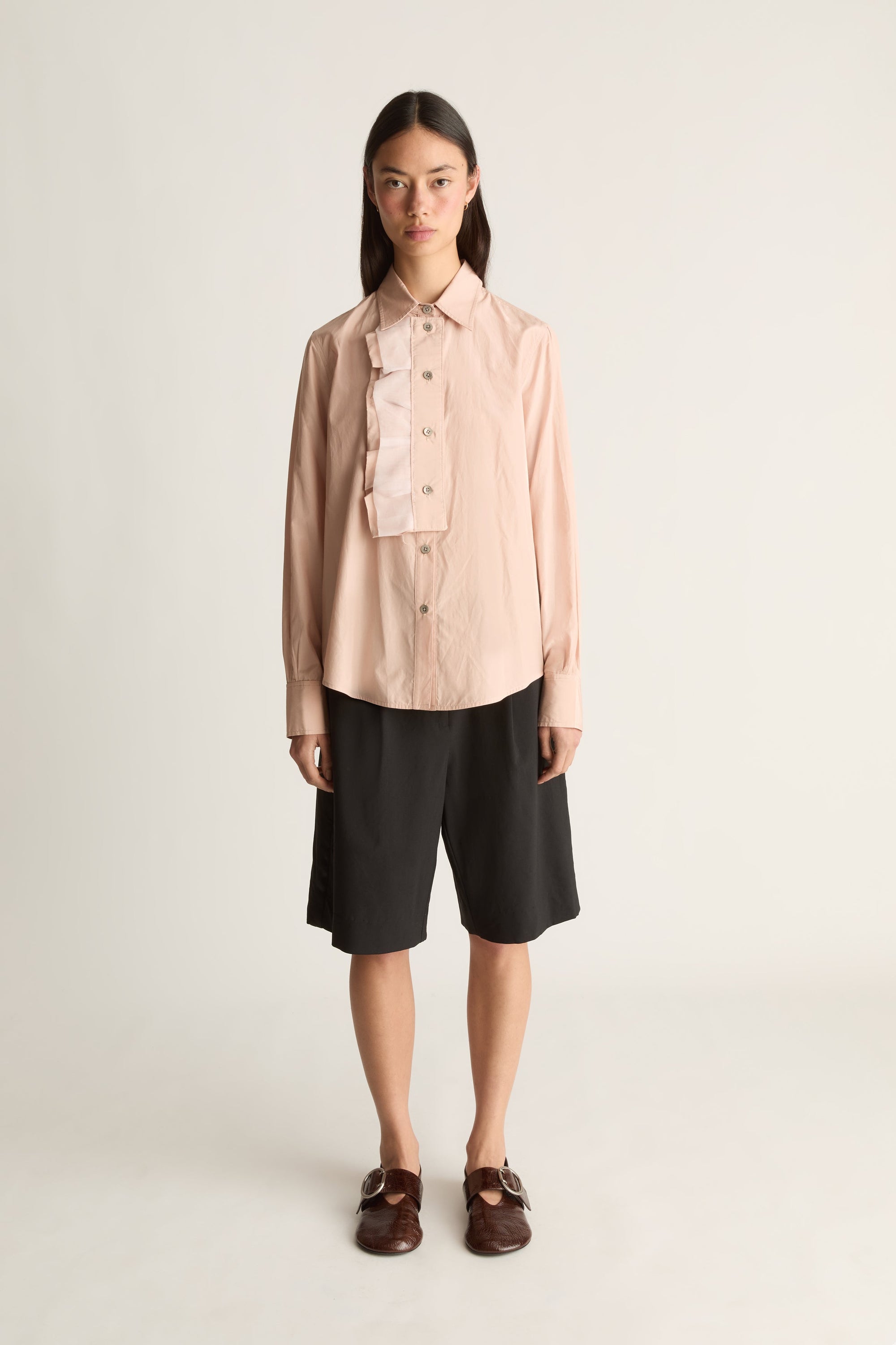 A model stands against a plain light gray studio background wearing a pale blush pink long-sleeve button-front shirt with a ruffled chest detail, black knee-length shorts, and dark brown loafers.
