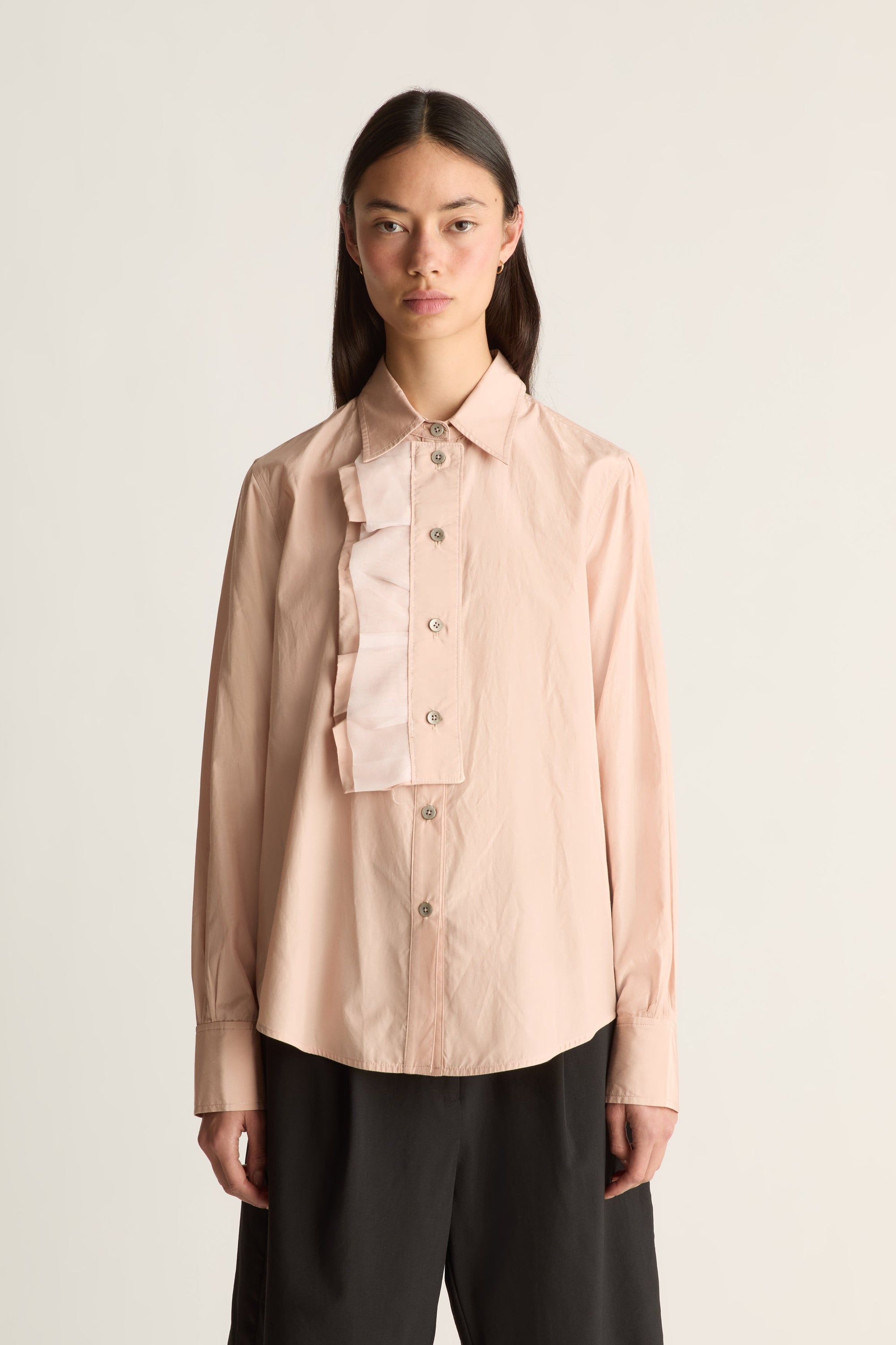 Close-up of a model wearing a pale blush long-sleeve button-up shirt with a layered ruffle collar detail, paired with black trousers, against a plain off-white studio background.